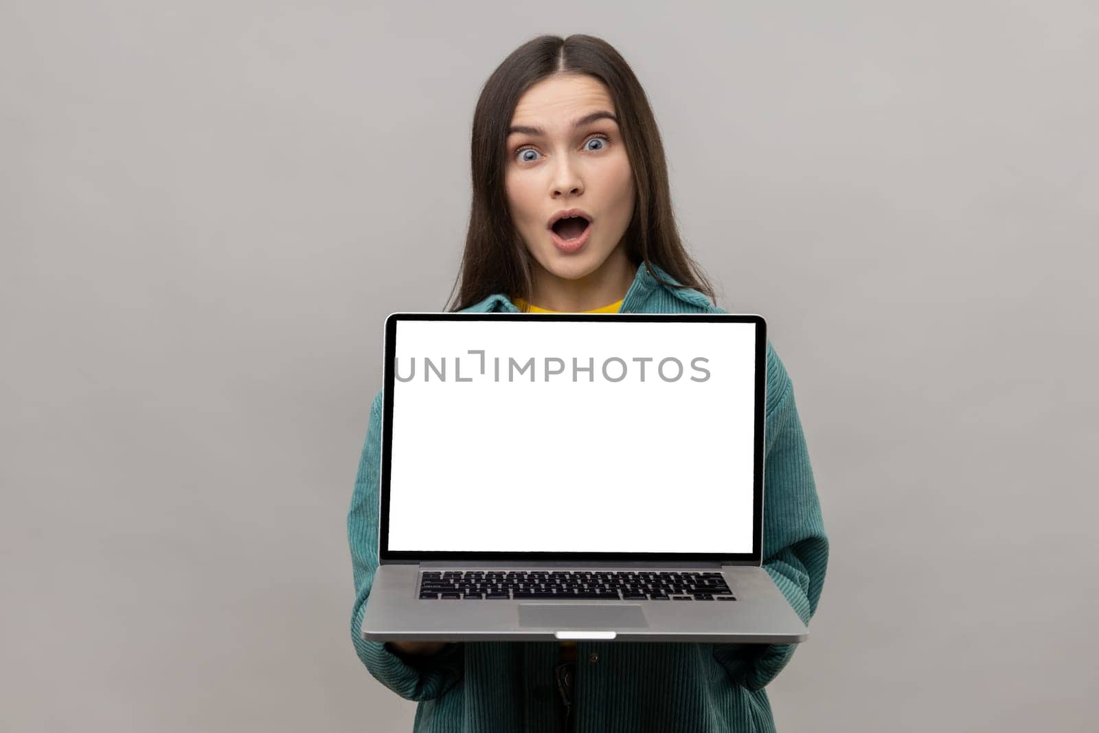 Surprised shocked woman standing showing laptop with white empty screen for promotion, looking at camera with open mouth, wearing casual style jacket. Indoor studio shot isolated on gray background.