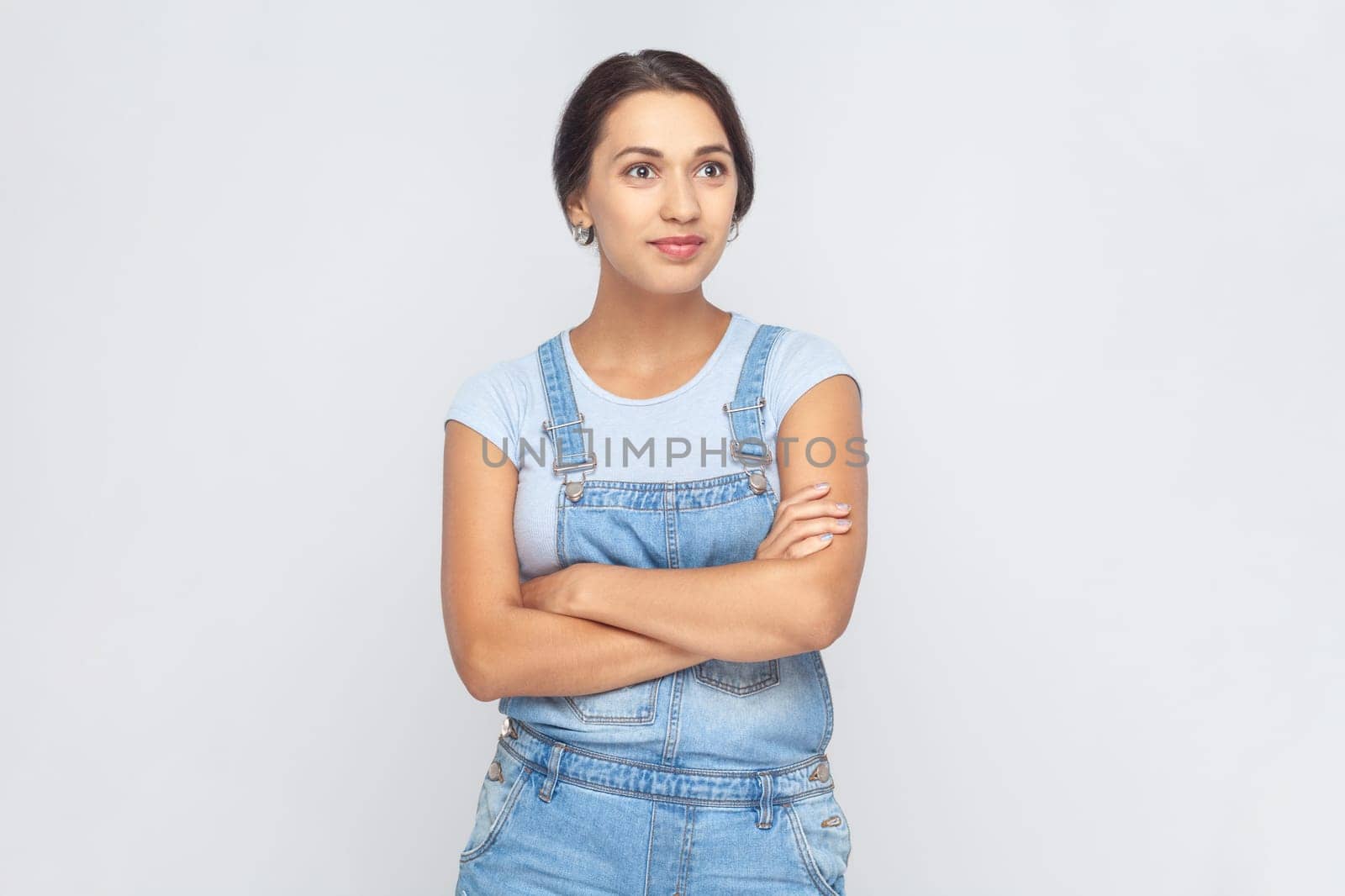 Portrait of calm smiling woman wearing denim overalls standing with crossed arms, looking at camera with confident facial expression. Indoor studio shot isolated on gray background.