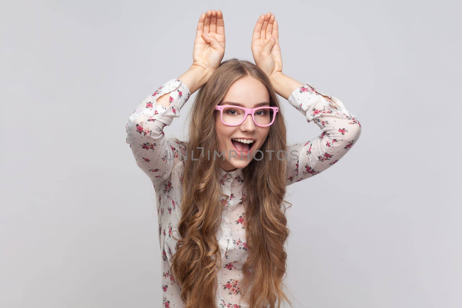 Portrait of funny woman in glasses with wavy blond hair showing bunny ears gesture and looking at camera with playful attractive smile. Indoor studio shot isolated on gray background.