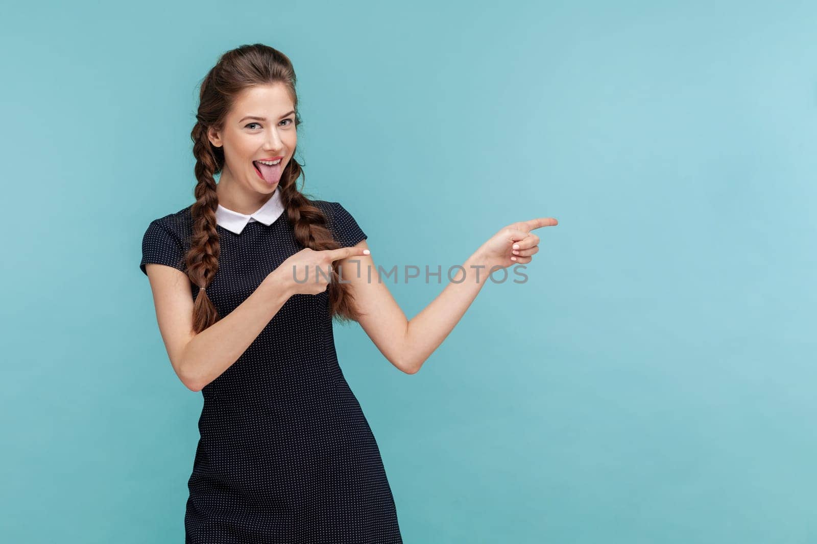 Portrait of cheerful lovely woman with braids sticks tongue, , indicates with both fore fingers at blank space, wearing black dress. woman Indoor studio shot isolated on blue background.