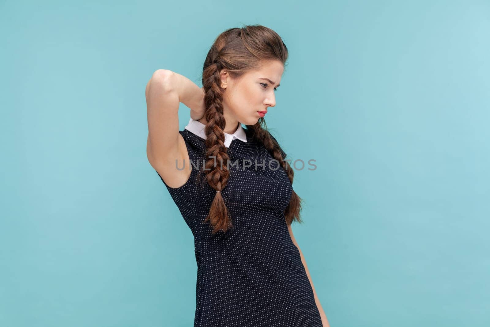 Portrait of unhappy sick woman with braids feels pain in neck, grimaces from pain, suffers without painkillers, wearing black dress. woman Indoor studio shot isolated on blue background.