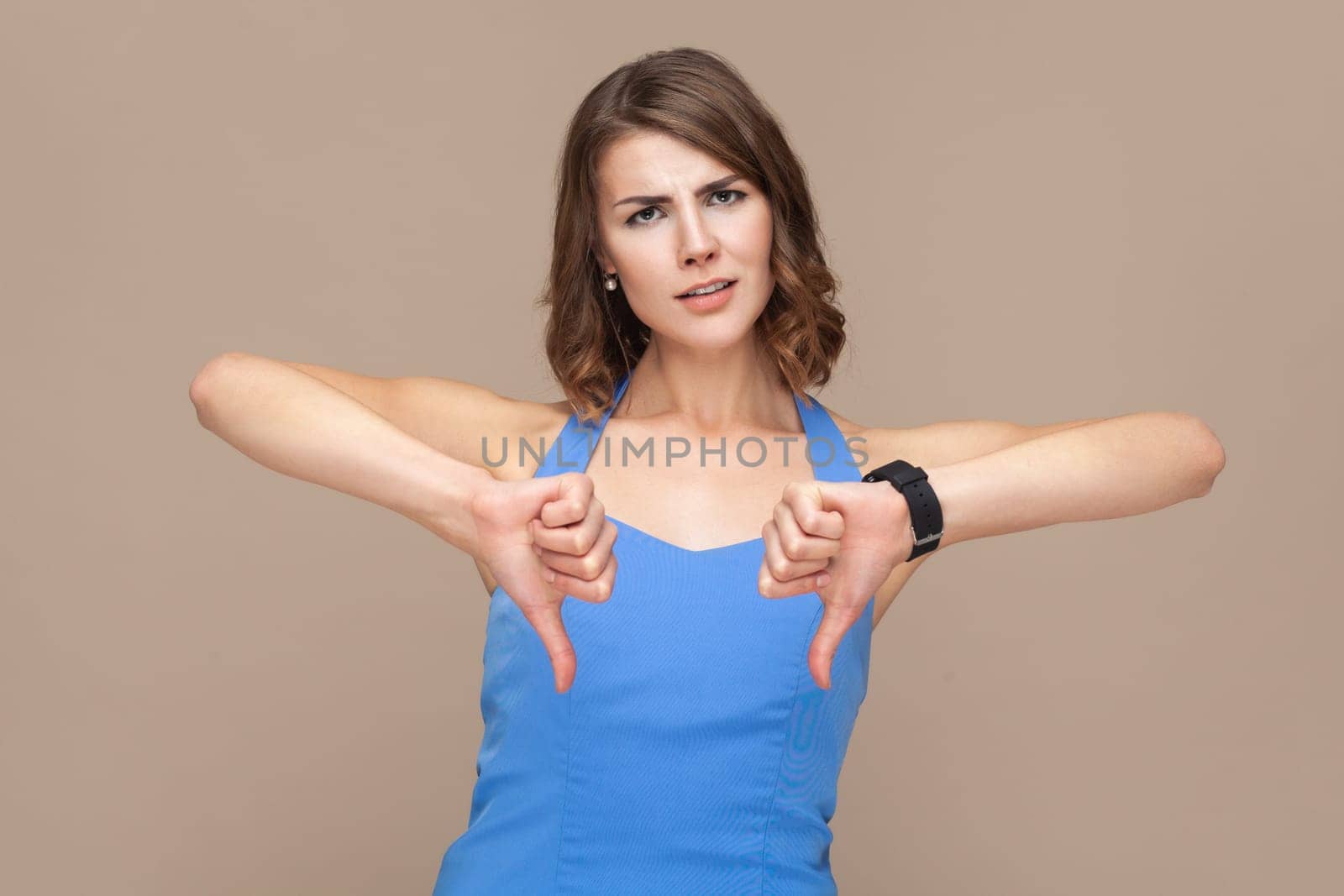 Attractive disappointed woman with wavy hair keeps thumb down, shows her dislike and disapproval as evaluates something, wearing blue dress. Indoor studio shot isolated on light brown background