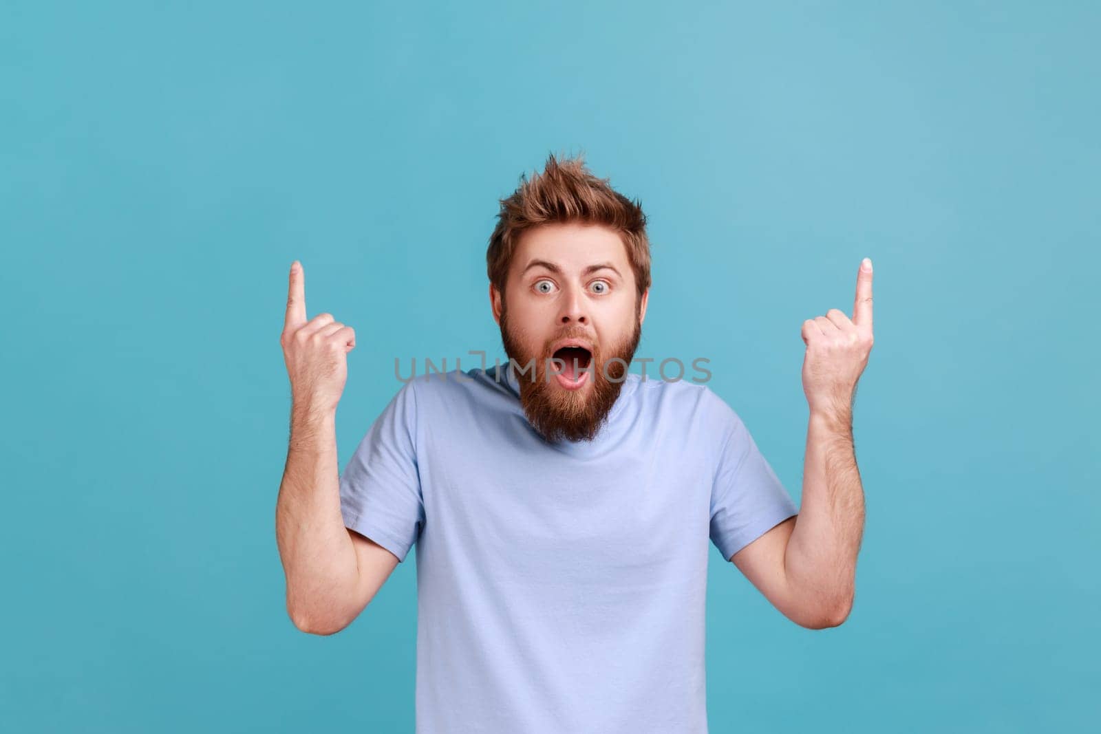 Wow, attention to advertising above. Portrait of bearded man pointing up at copy space and looking surprised, showing blank wall for idea presentation. Indoor studio shot isolated on blue background.