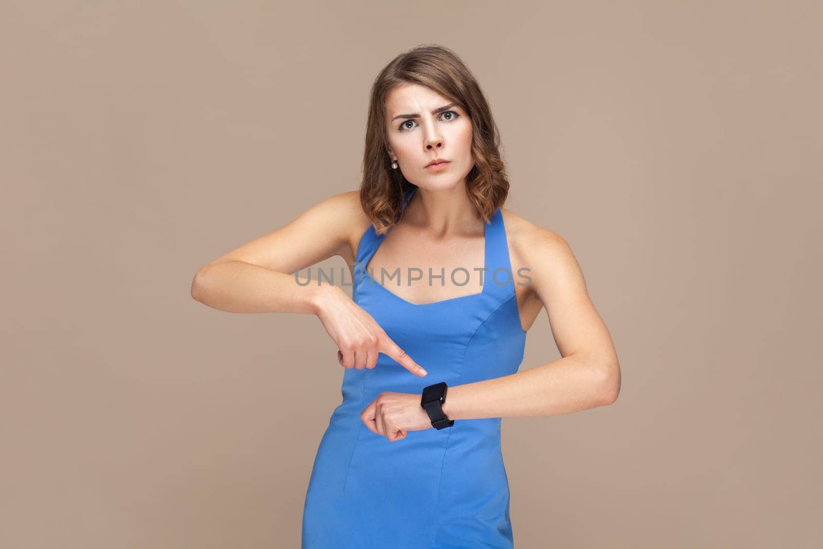 You are late. Portrait of serious woman with wavy hair standing, looking at camera and pointing at her smart watch, wearing blue dress. Indoor studio shot isolated on light brown background.