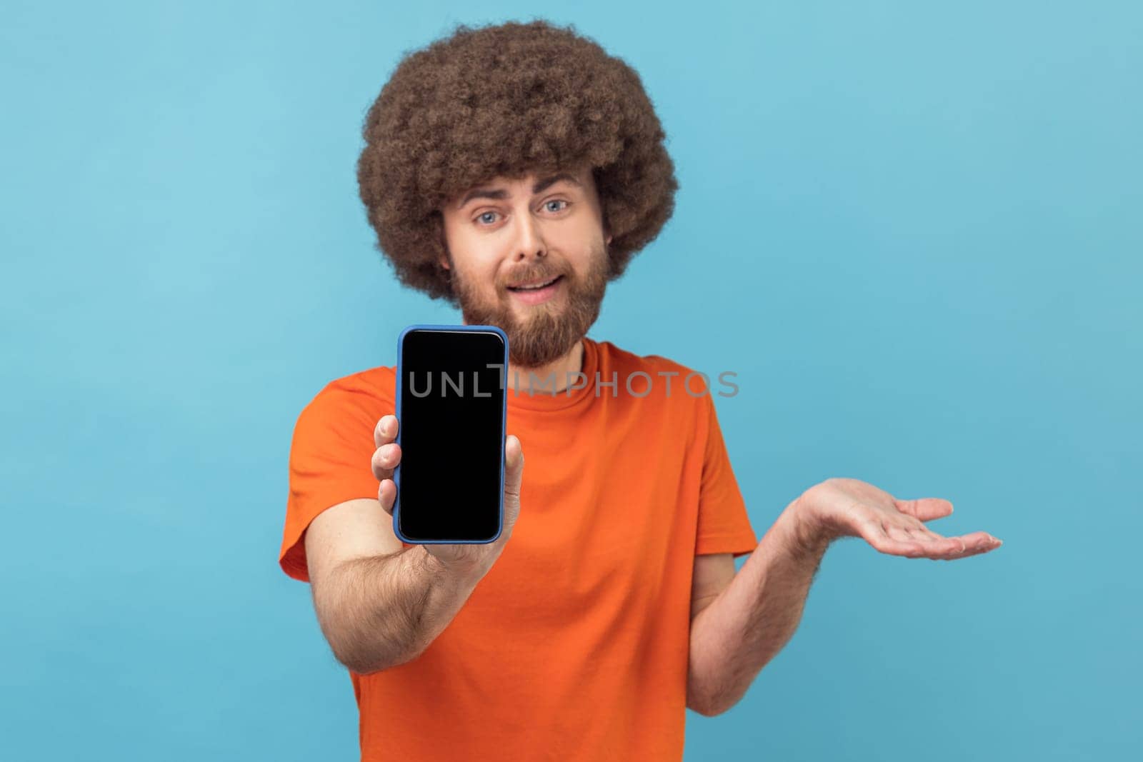 Portrait of confused man with Afro hairstyle wearing orange T-shirt showing cell phone with blank black screen, presenting area for advertisement. Indoor studio shot isolated on blue background.