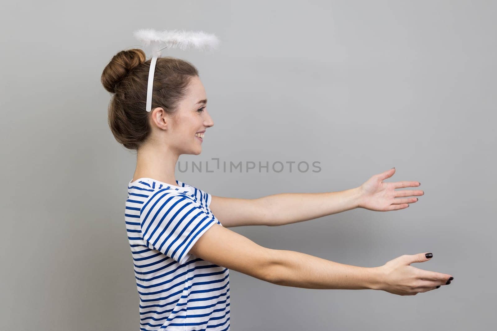 Side view of woman wearing striped T-shirt and with nimbus over her head stretching arms with kind friendly smile, going to embrace, share love. Indoor studio shot isolated on gray background
