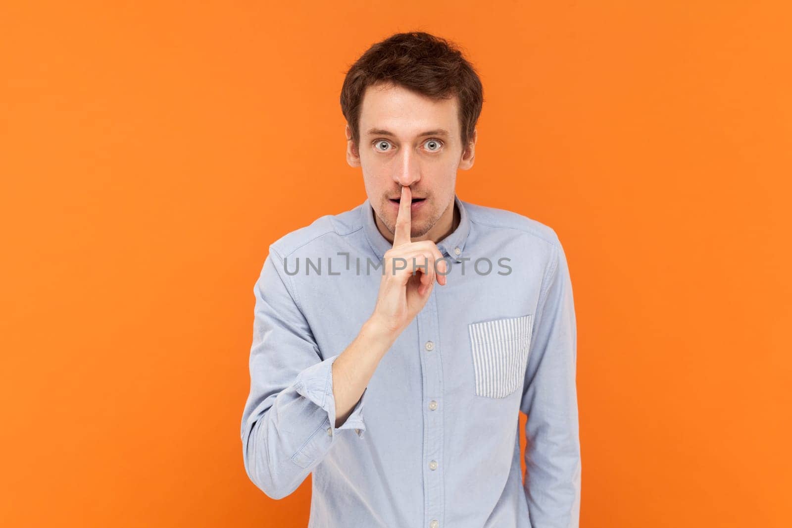 Serious young adult man standing with finger near lips, asking to keep silent, looking at camera with bossy expression, wearing light blue shirt. Indoor studio shot isolated on orange background.