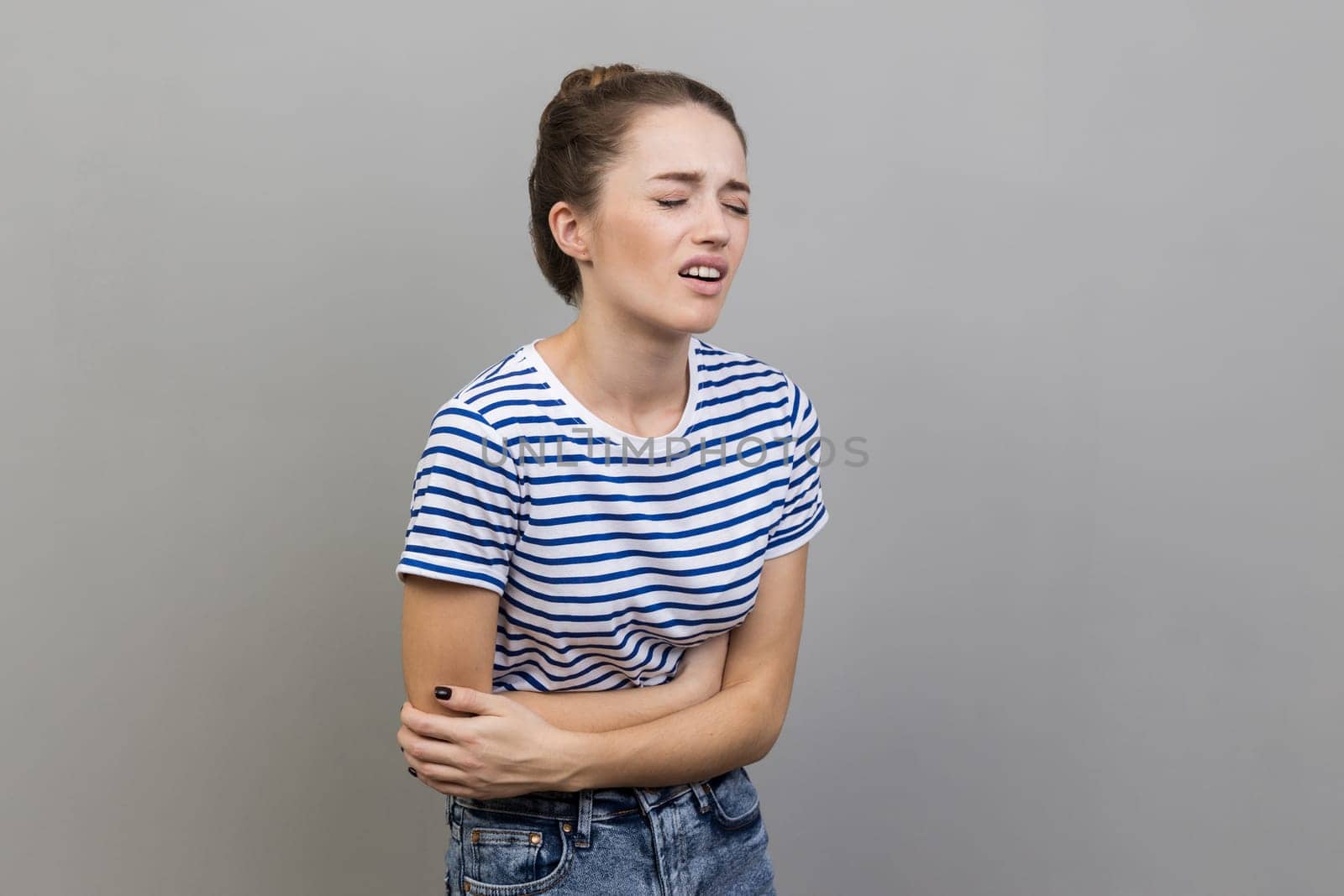 Portrait of unhealthy sick woman wearing striped T-shirt standing and touching her painful belly, frowning face from terrible pain. Indoor studio shot isolated on gray background.