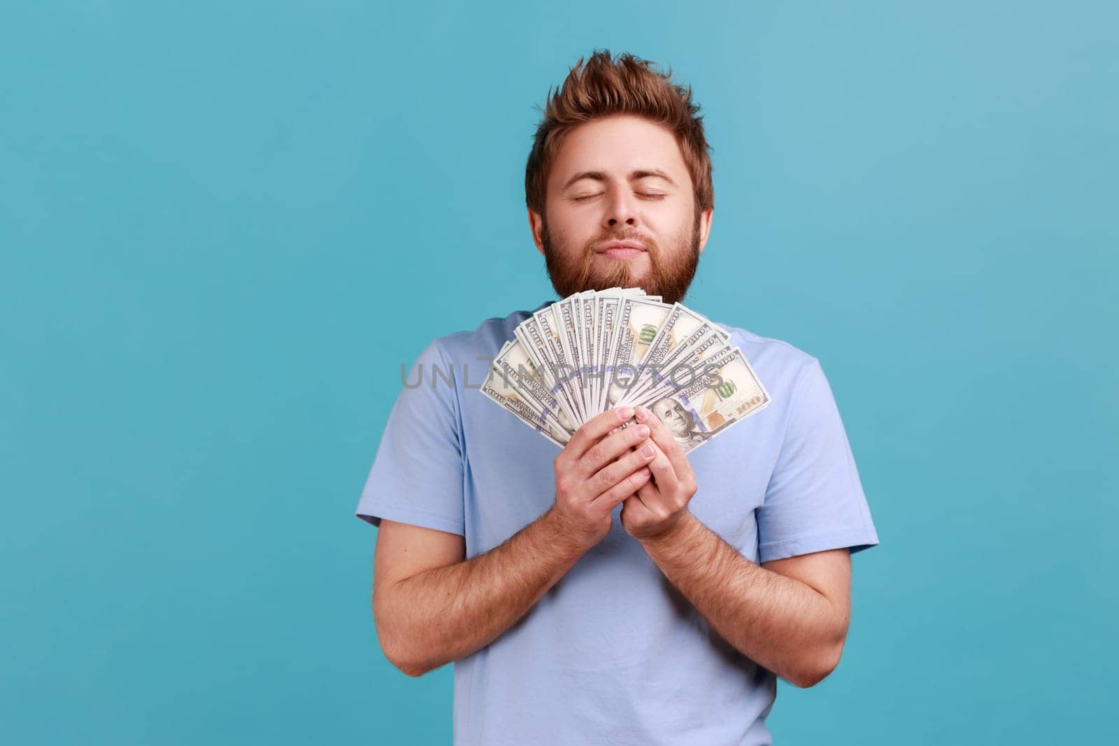 Portrait of satisfied bearded man smelling earned dollar banknotes, enjoying success and big profit, wealthy life, greedy for money. Indoor studio shot isolated on blue background.