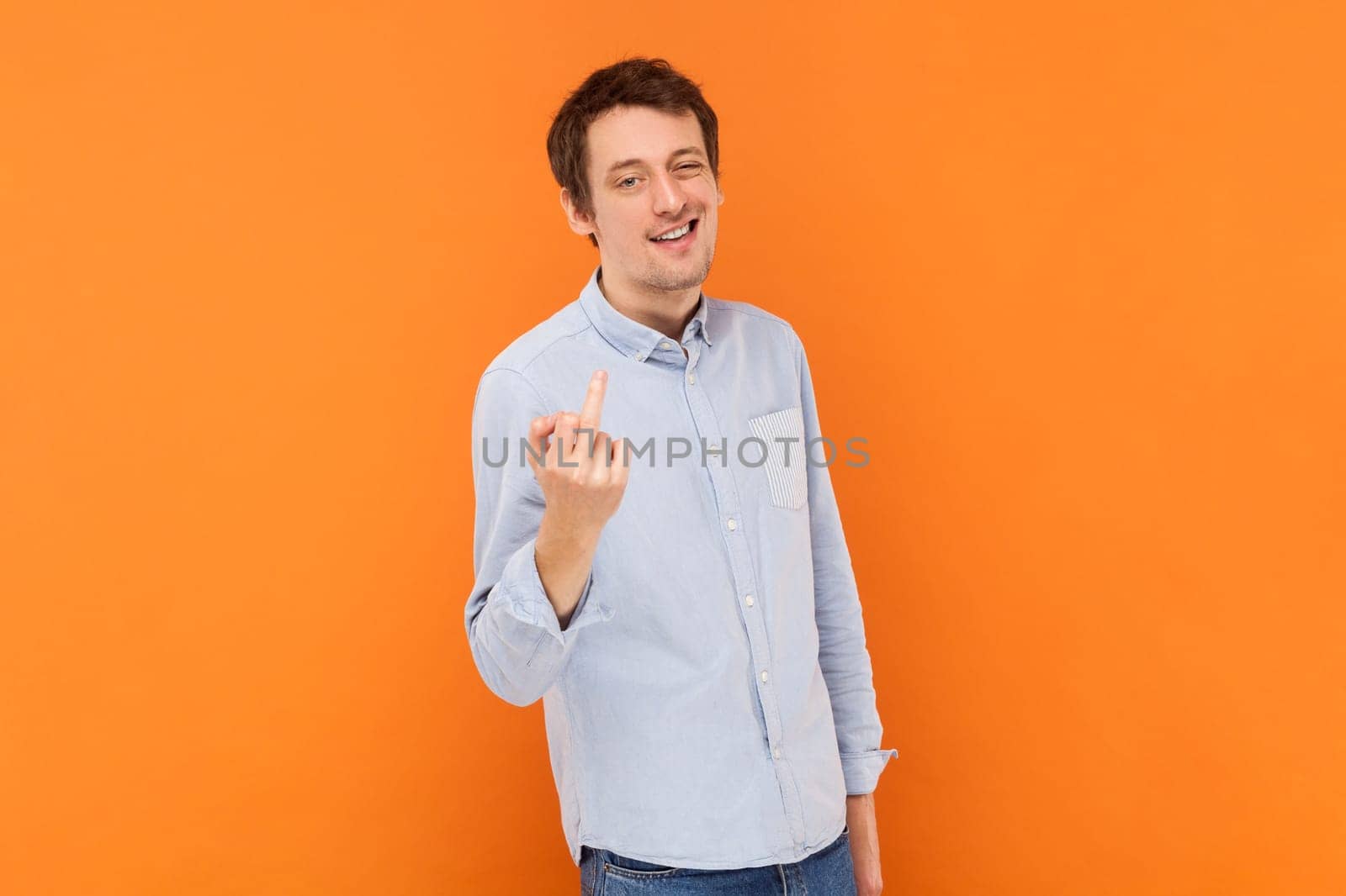 Portrait of serious vulgar selfish man showing middle finger, impolite gesture, disrespects someone, looks rude at camera, wearing light blue shirt. Indoor studio shot isolated on orange background.