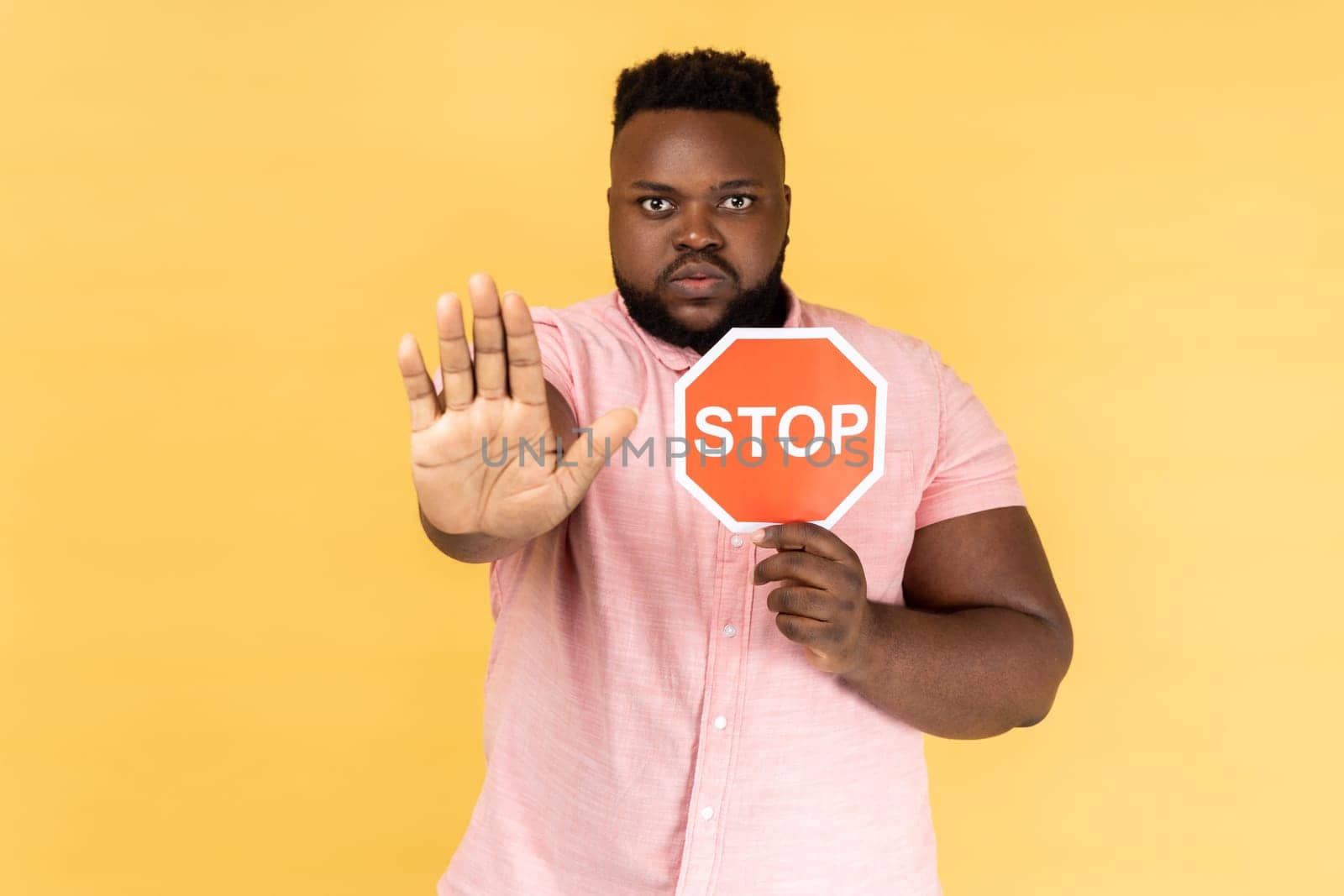 Portrait of man wearing pink shirt holding red stop sign, looking at camera with ban palm gesture, has strict expression, prohibition. Indoor studio shot isolated on yellow background.