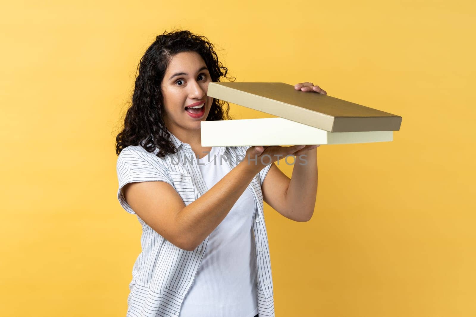 Portrait of woman with dark wavy hair holding in hands half opens wrapped present box, looking at camera with excitement and surprise. Indoor studio shot isolated on yellow background.