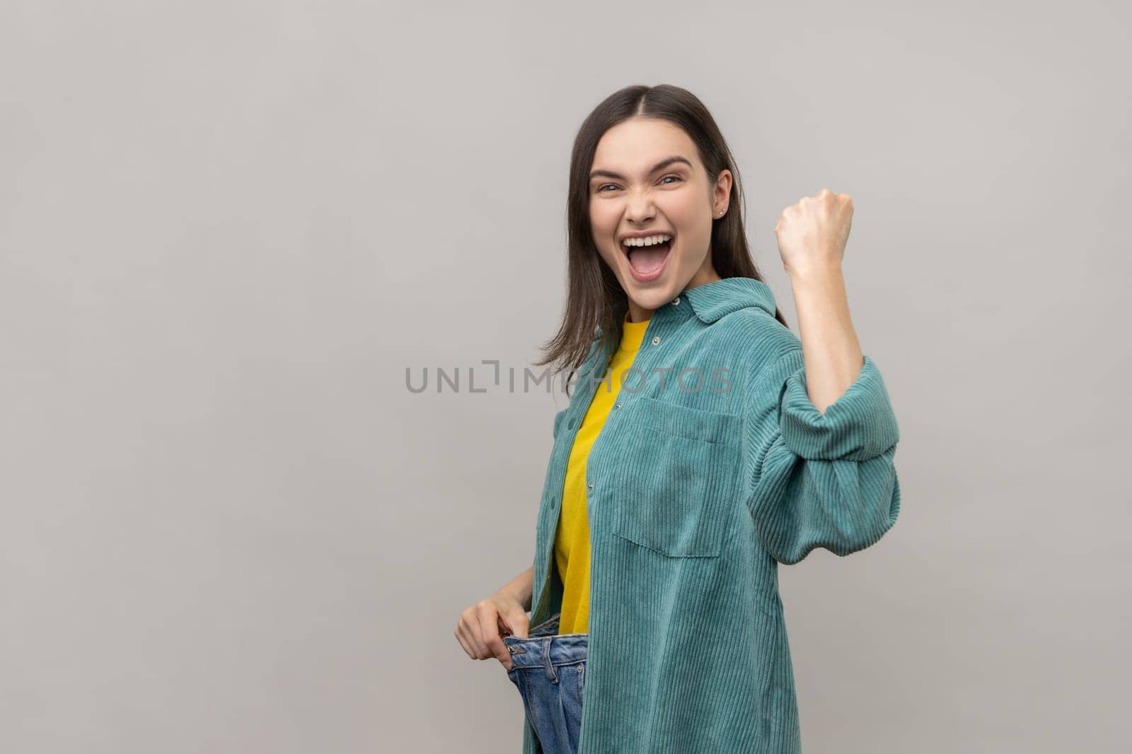Attractive happy woman have willpower to loose weight, showing slim waist and clenched fist, celebrating, diet conception, wearing casual style jacket. Indoor studio shot isolated on gray background.