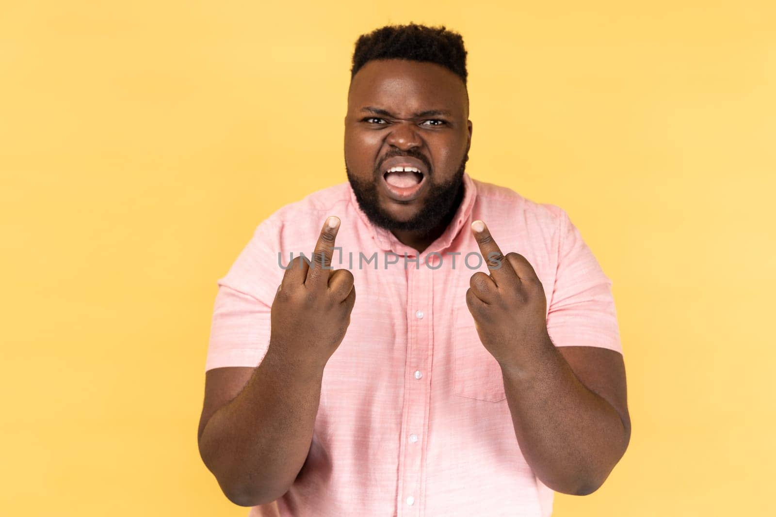 Portrait of impolite aggressive man wearing pink shirt showing middle fingers and asking to get off expressing negativity, disrespectful behaviour. Indoor studio shot isolated on yellow background.