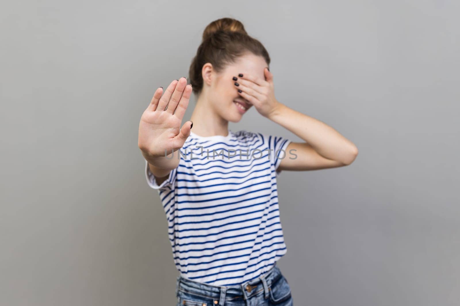 Portrait of afraid beautiful woman wearing striped T-shirt closing eyes with palm and showing stop hand gesture, turning face, does not want to see. Indoor studio shot isolated on gray background.