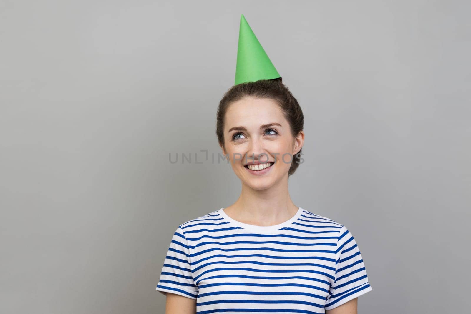 Portrait of delighted smiling dreamy woman wearing striped T-shirt and party cone looking away with satisfied smile, celebrating birthday. Indoor studio shot isolated on gray background.