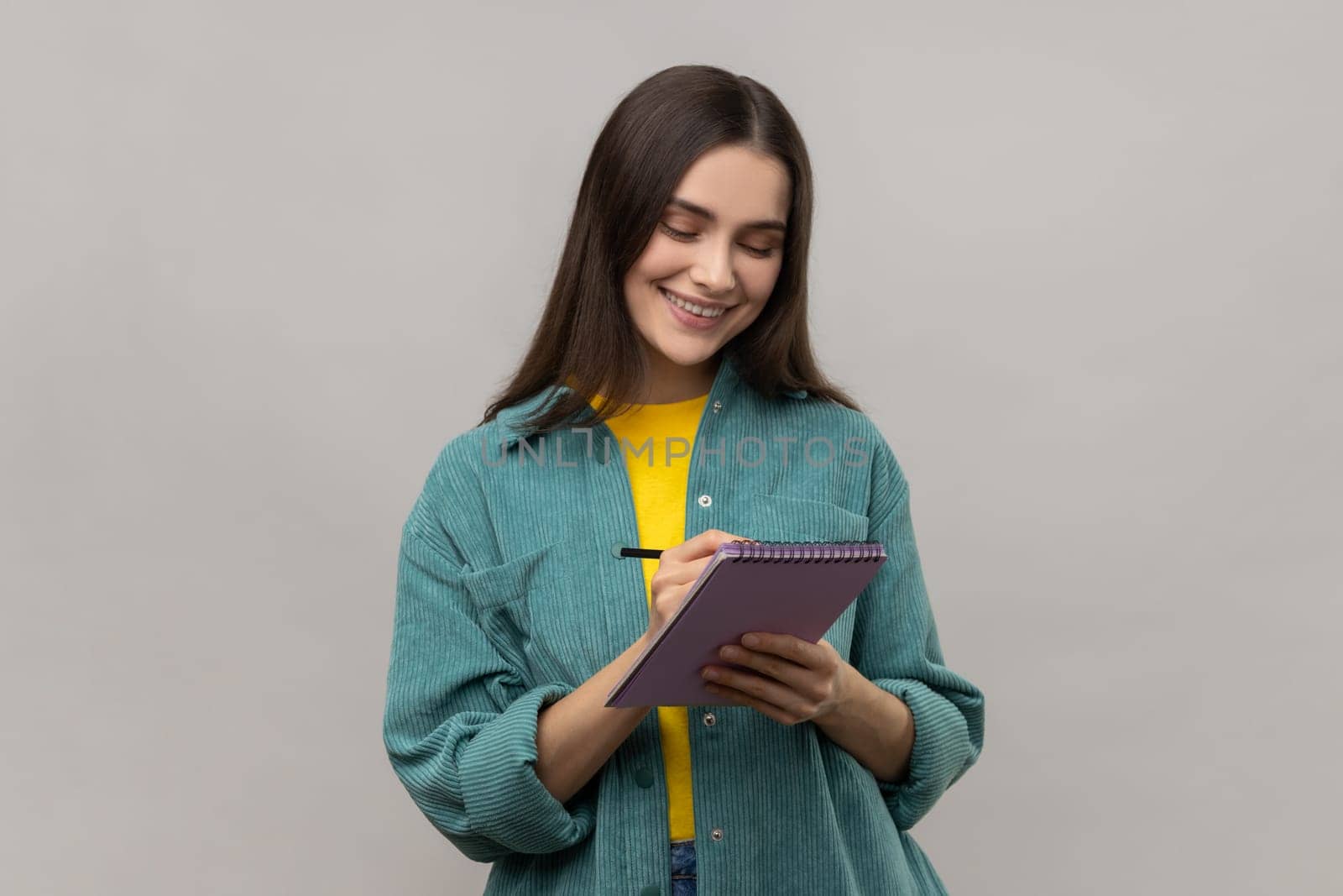 Portrait of happy inspired woman smiling and writing down interesting idea in notebook, making plans, to-do list, wearing casual style jacket. Indoor studio shot isolated on gray background.