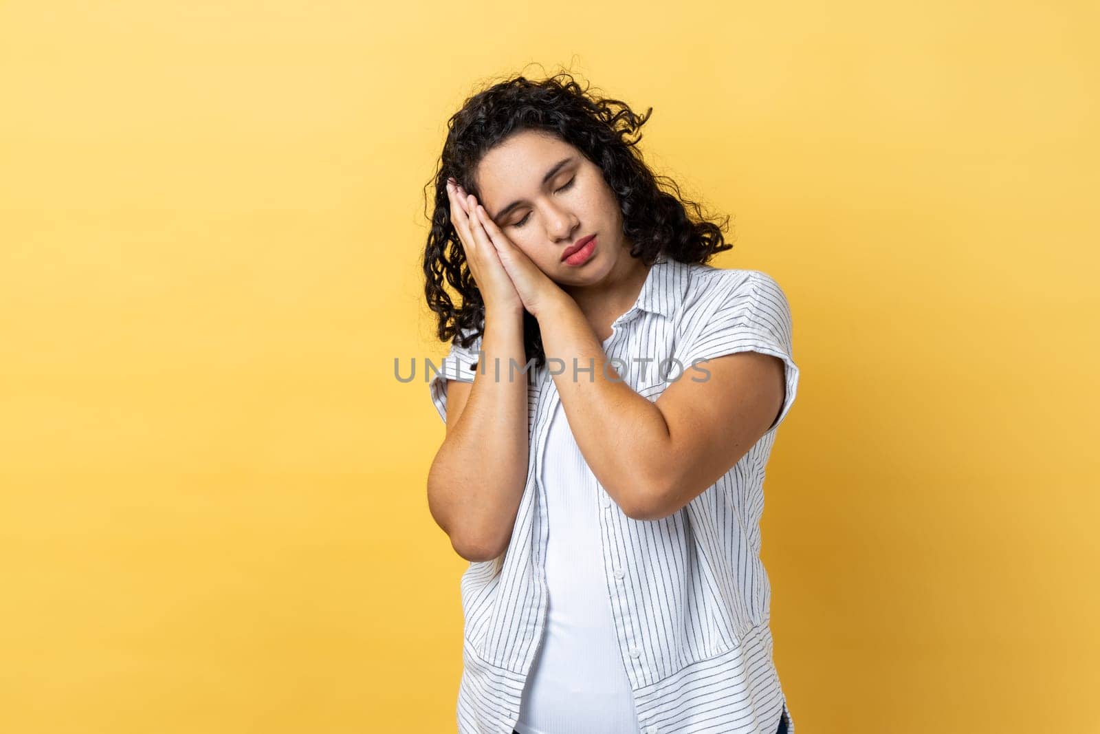 Portrait of exhausted tired beautiful woman with dark wavy hair sleeping laying down on palms, having comfortable nap and resting. Indoor studio shot isolated on yellow background.