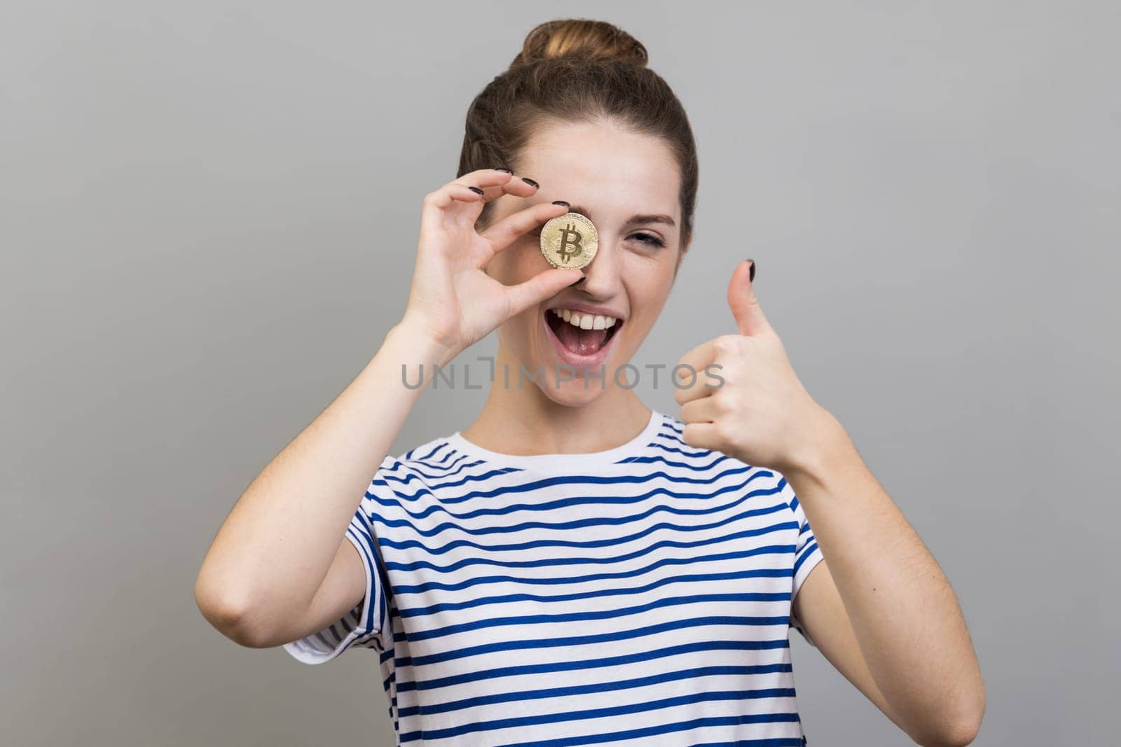 Portrait of amazed woman wearing striped T-shirt covering eye with golden bitcoin, looking at camera through btc coin and showing thumb up. Indoor studio shot isolated on gray background.