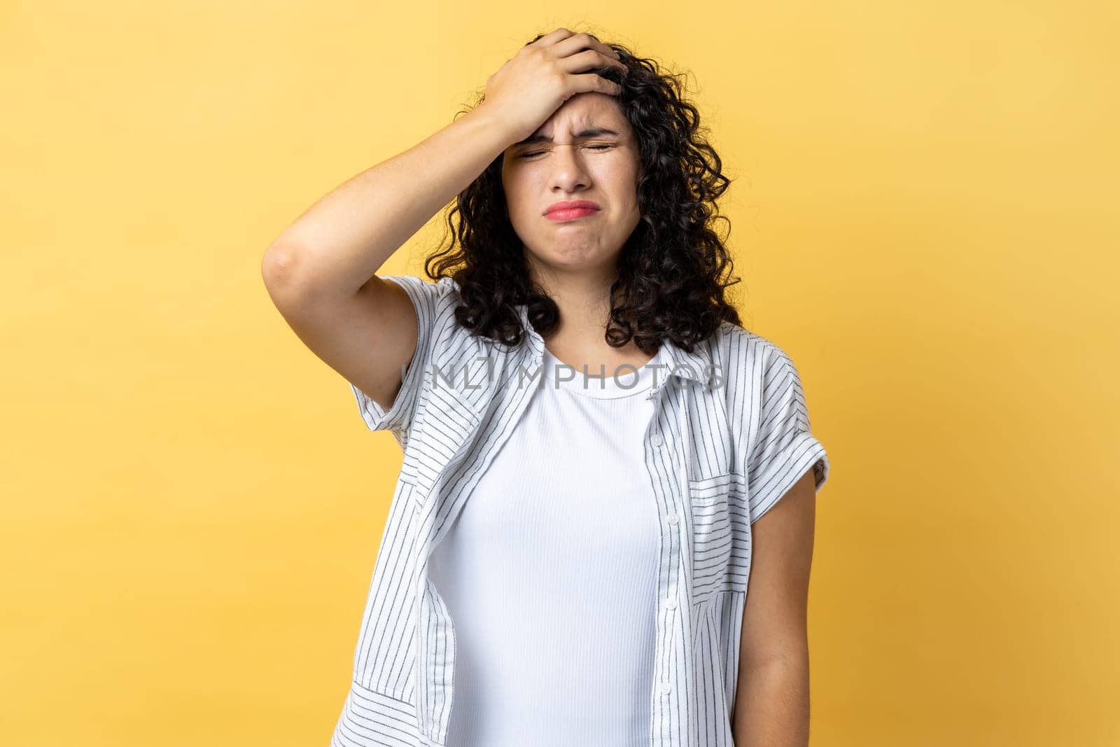 Portrait of woman with dark wavy hair standing with facepalm gesture, blaming herself, feeling sorrow regret because of bad memory. Indoor studio shot isolated on yellow background.