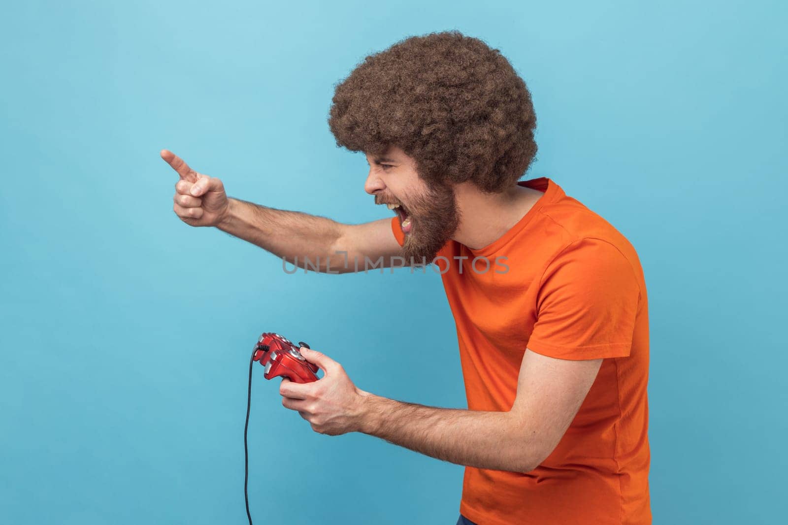 Side view of excited man with Afro hairstyle in orange T-shirt holding in hands red gamepad joystick, grimacing playing video games, raised finger up. Indoor studio shot isolated on blue background.