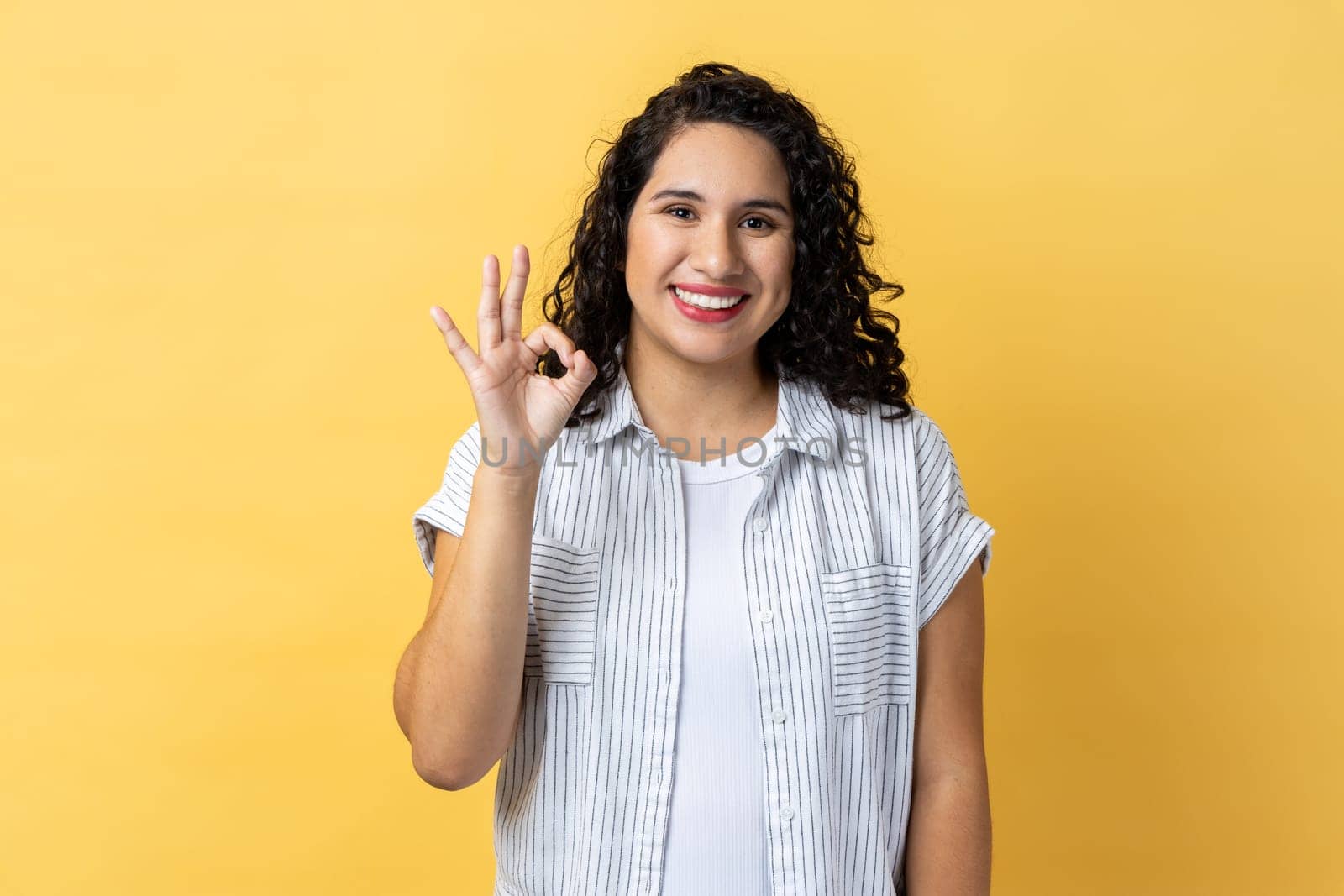 Portrait of smiling satisfied positive optimistic beautiful woman with dark wavy hair looking at camera showing ok sign gesture. Indoor studio shot isolated on yellow background.