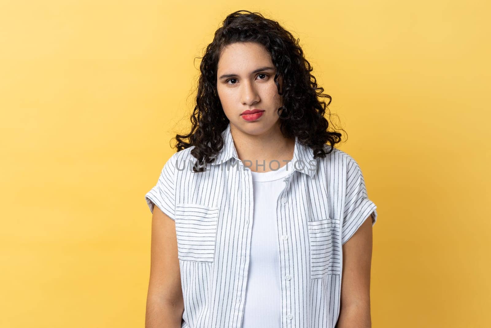 Portrait of sad desperate woman with dark wavy hair standing looking at camera, being in bad mood, suffering depression. Indoor studio shot isolated on yellow background.