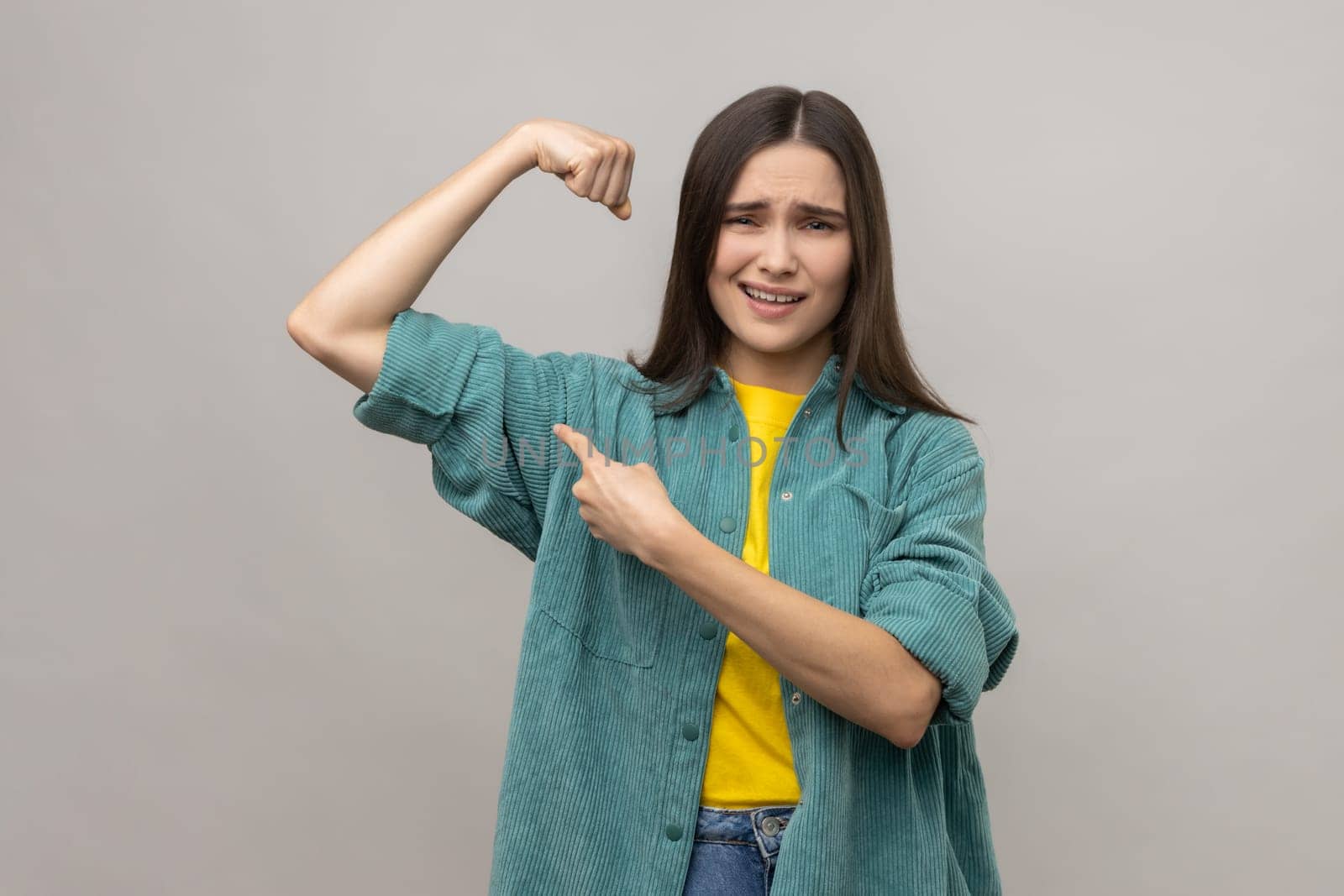 Look how strong I am. Confident independent woman raising hand pointing biceps, feeling power and energy to achieve goal, wearing casual style jacket. Indoor studio shot isolated on gray background.