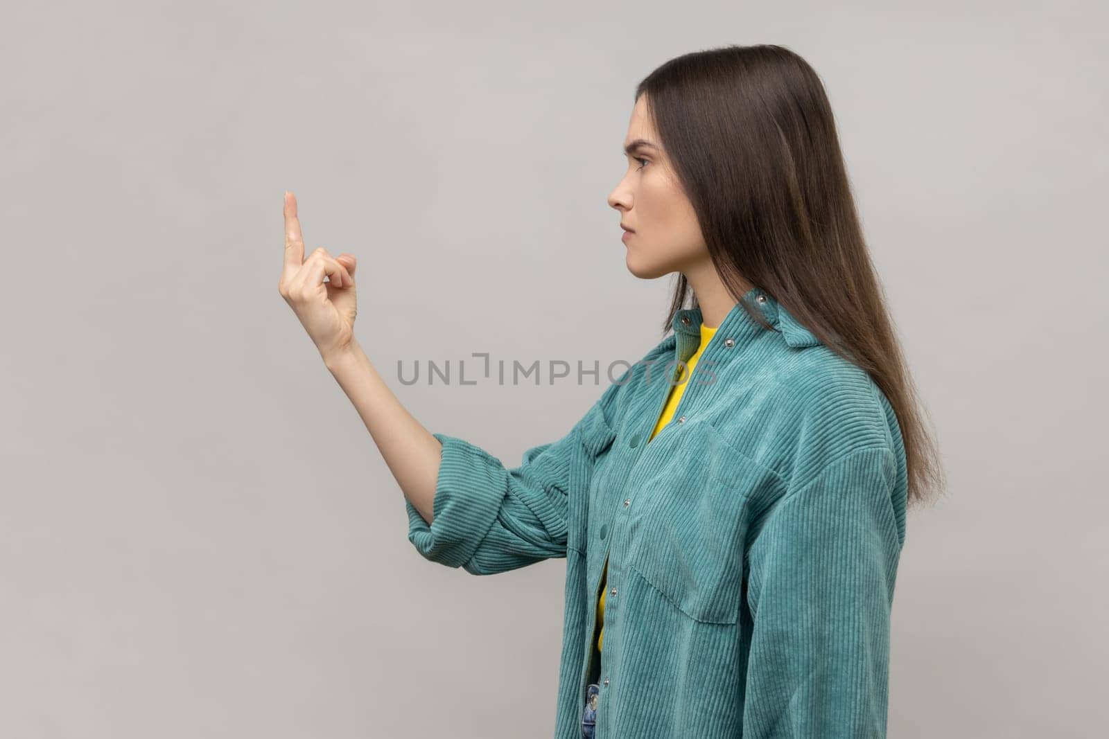 Side view of dark haired woman rejecting communication, showing middle finger to express disrespect and hate vulgar gesture, wearing casual style jacket. Indoor studio shot isolated on gray background