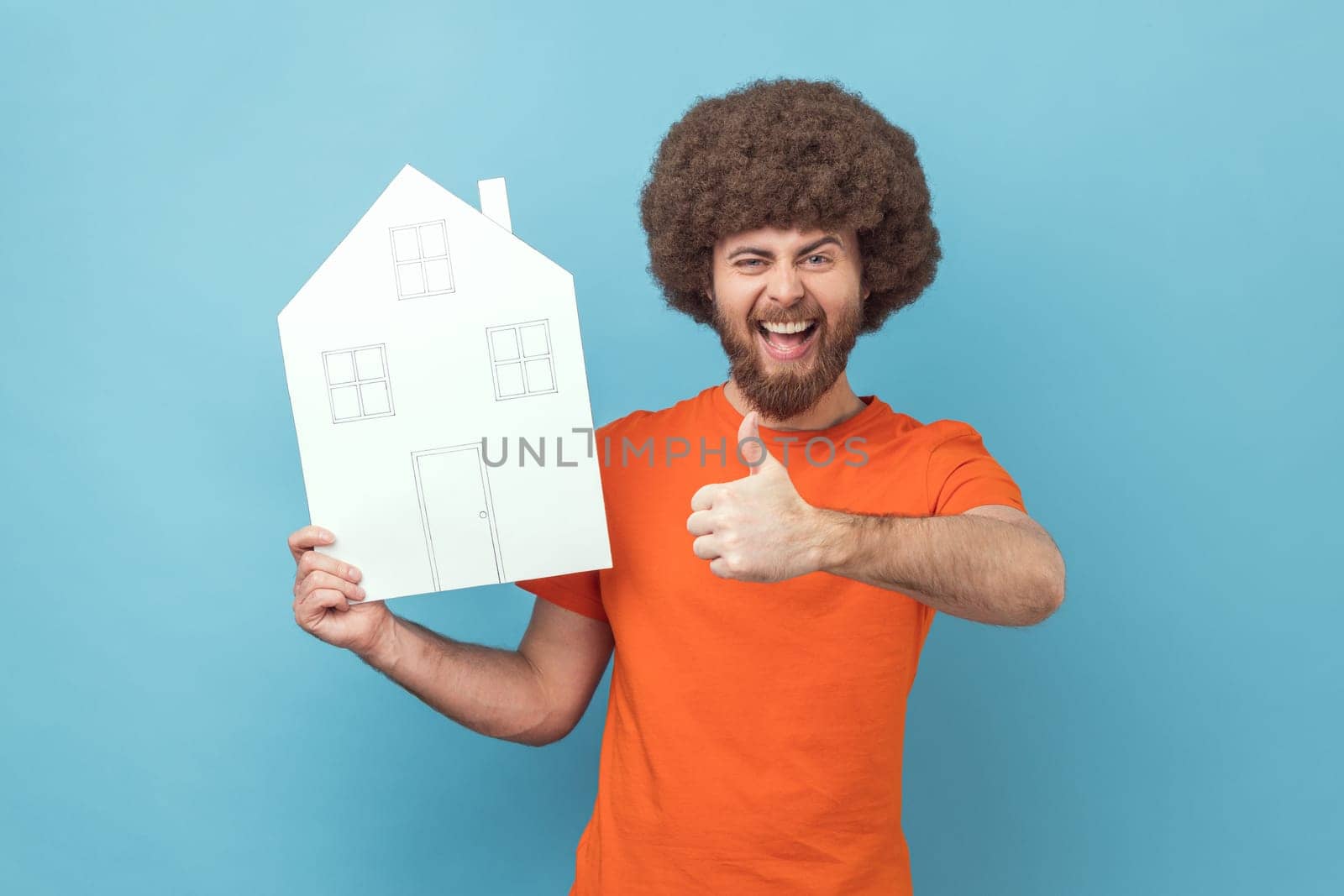 Portrait of excited positive man with Afro hairstyle wearing orange T-shirt holding paper house, real estate agency, showing thumb up. Indoor studio shot isolated on blue background.