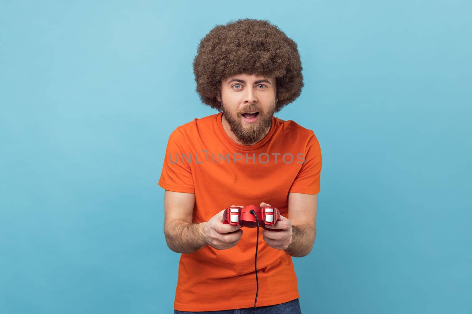 Portrait of concentrated excited man with Afro hairstyle grimacing holding joystick, playing video games online, virtual competition. Indoor studio shot isolated on blue background.