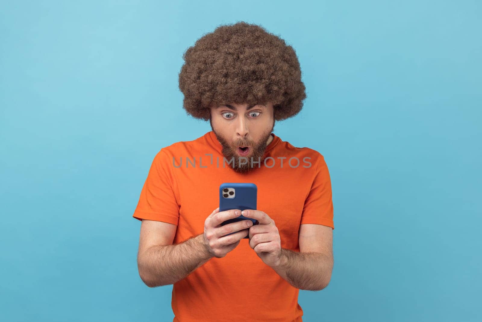 Portrait of astonished man with Afro hairstyle wearing orange T-shirt reading post on social network using cell phone, chatting looking surprised. Indoor studio shot isolated on blue background.