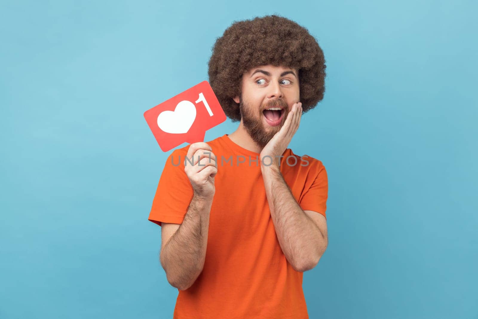 Portrait of cute surprised man with Afro hairstyle wearing orange T-shirt holding social media heart Like button, emoji counter, follower notification. Indoor studio shot isolated on blue background.