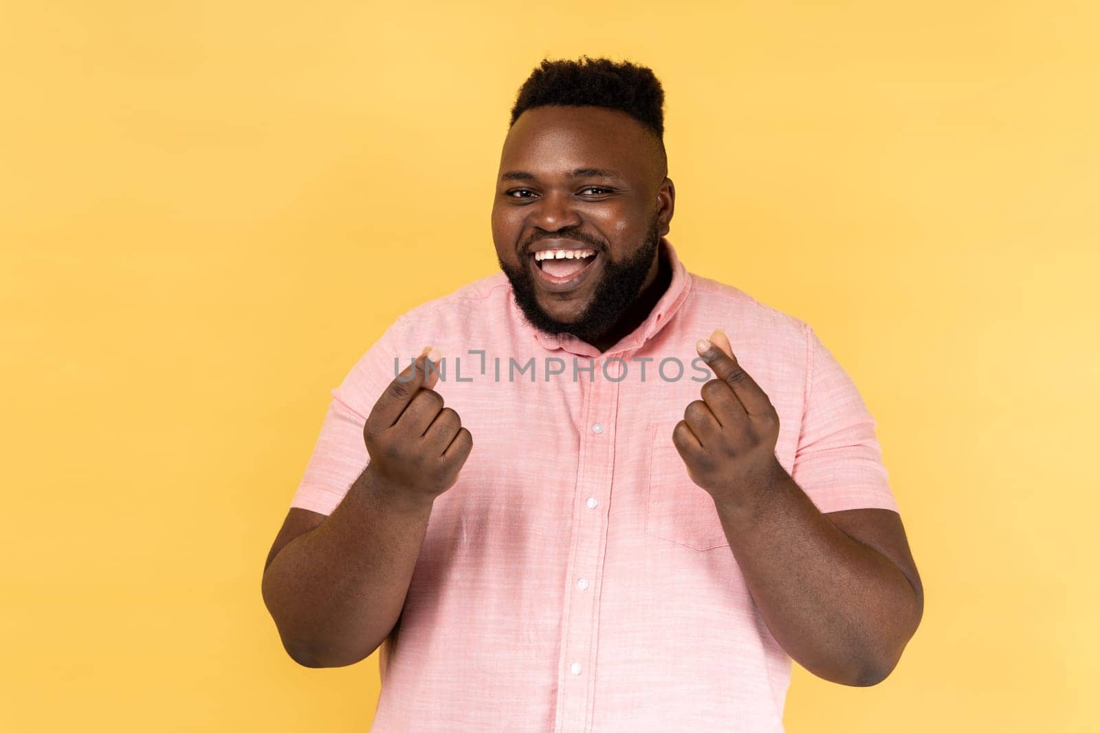 Portrait of smiling bearded man wearing pink shirt makes money gesture, rubs fingers, looking at camera with glad expression. Indoor studio shot isolated on yellow background.