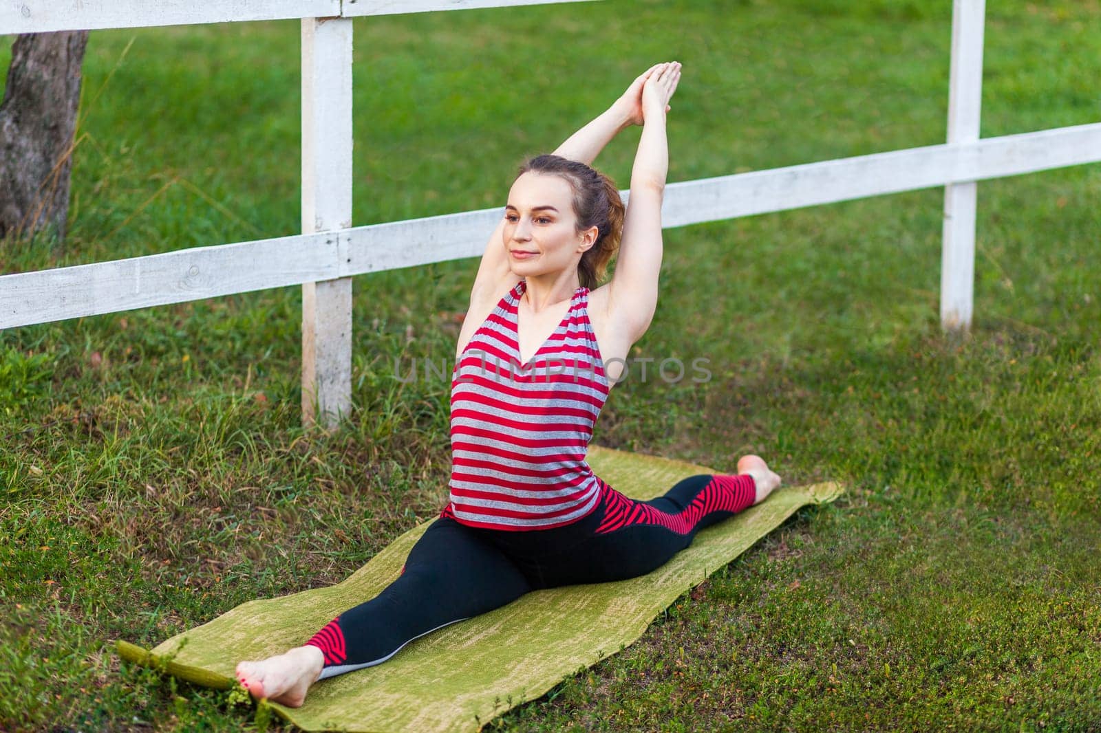 Full length portrait of young athletic yogi attractive woman practicing yoga in the park, doing Monkey God exercise, splits, Hanumanasana pose, working out, wearing sportswear.