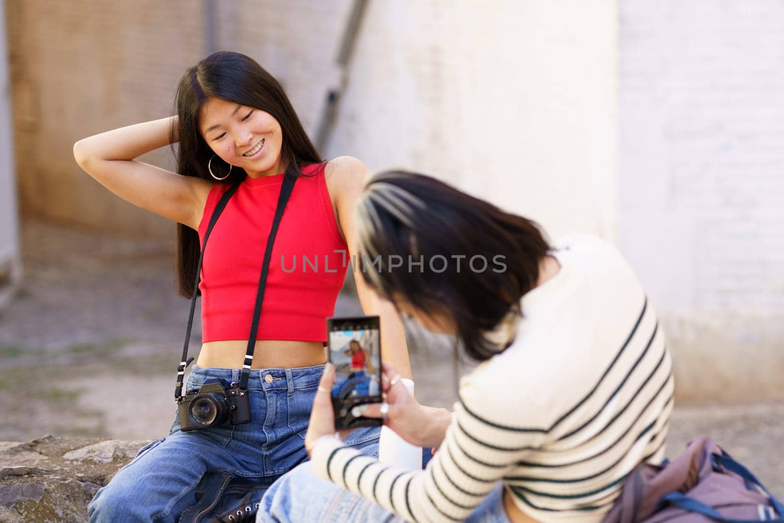 Positive young ethnic woman in red top and jeans touching hair while posing for photo in front of girlfriend in striped sweater during sightseeing