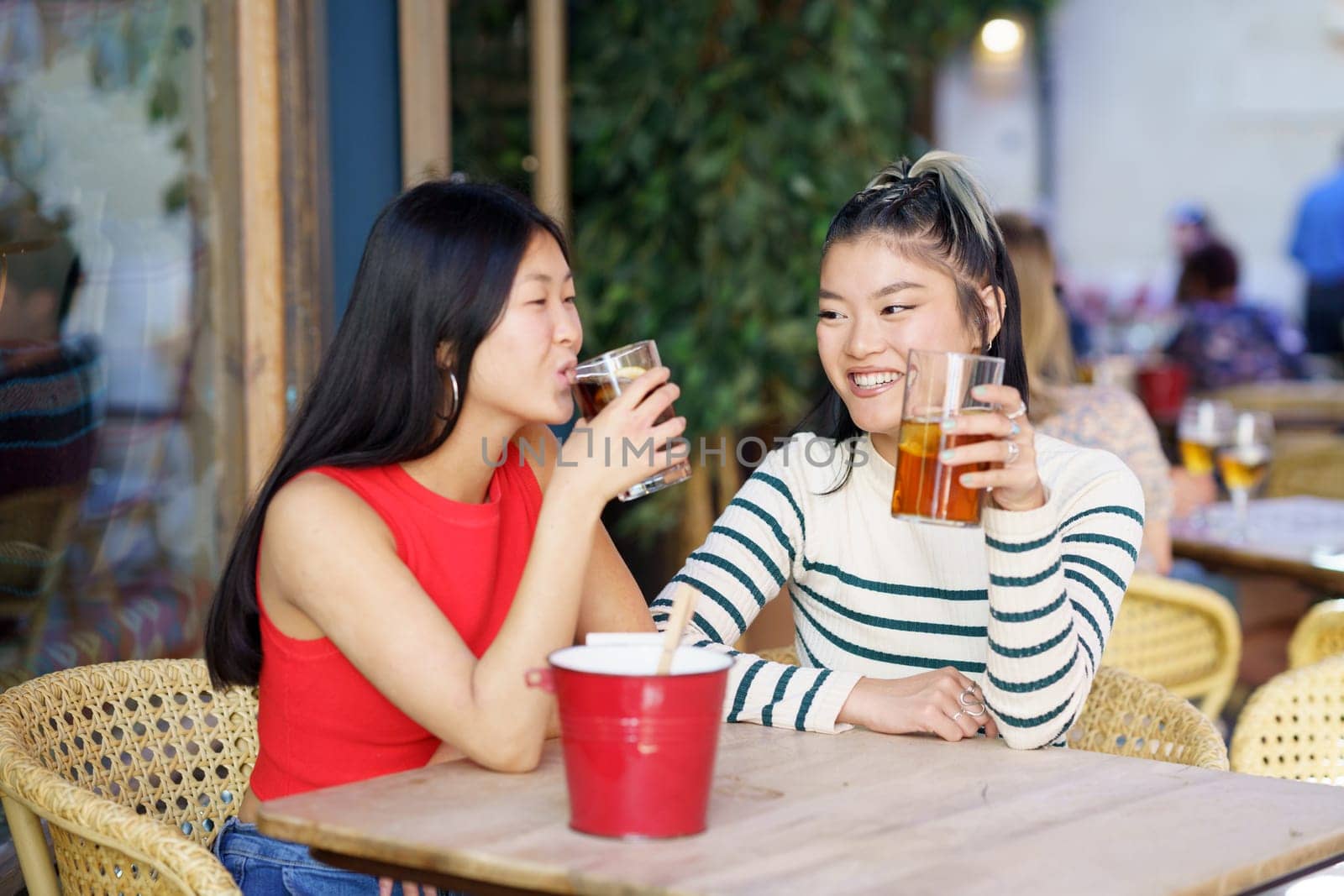 Cheerful young Asian female friends in casual wear sitting at wooden table and chatting while drinking refreshing cold tea during meeting in cafe