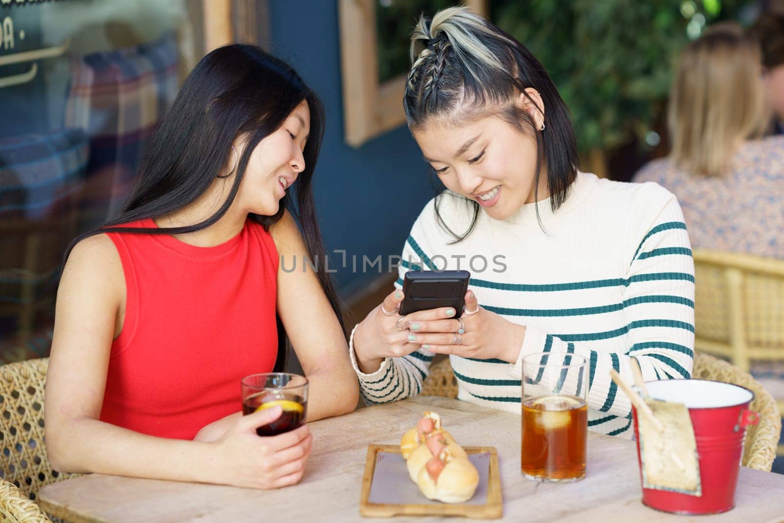 Content young Asian female friends in casual outfits, smiling and taking photo of delicious meal on smartphone while gathering at wooden table with glasses of drinks in cafe