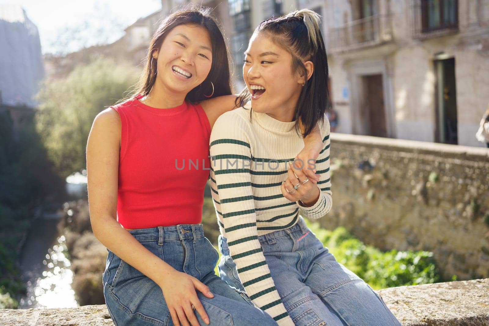 Happy young Asian female putting hand on shoulder of friend in casual clothes smiling and looking at camera while sitting on stone bench in Granada