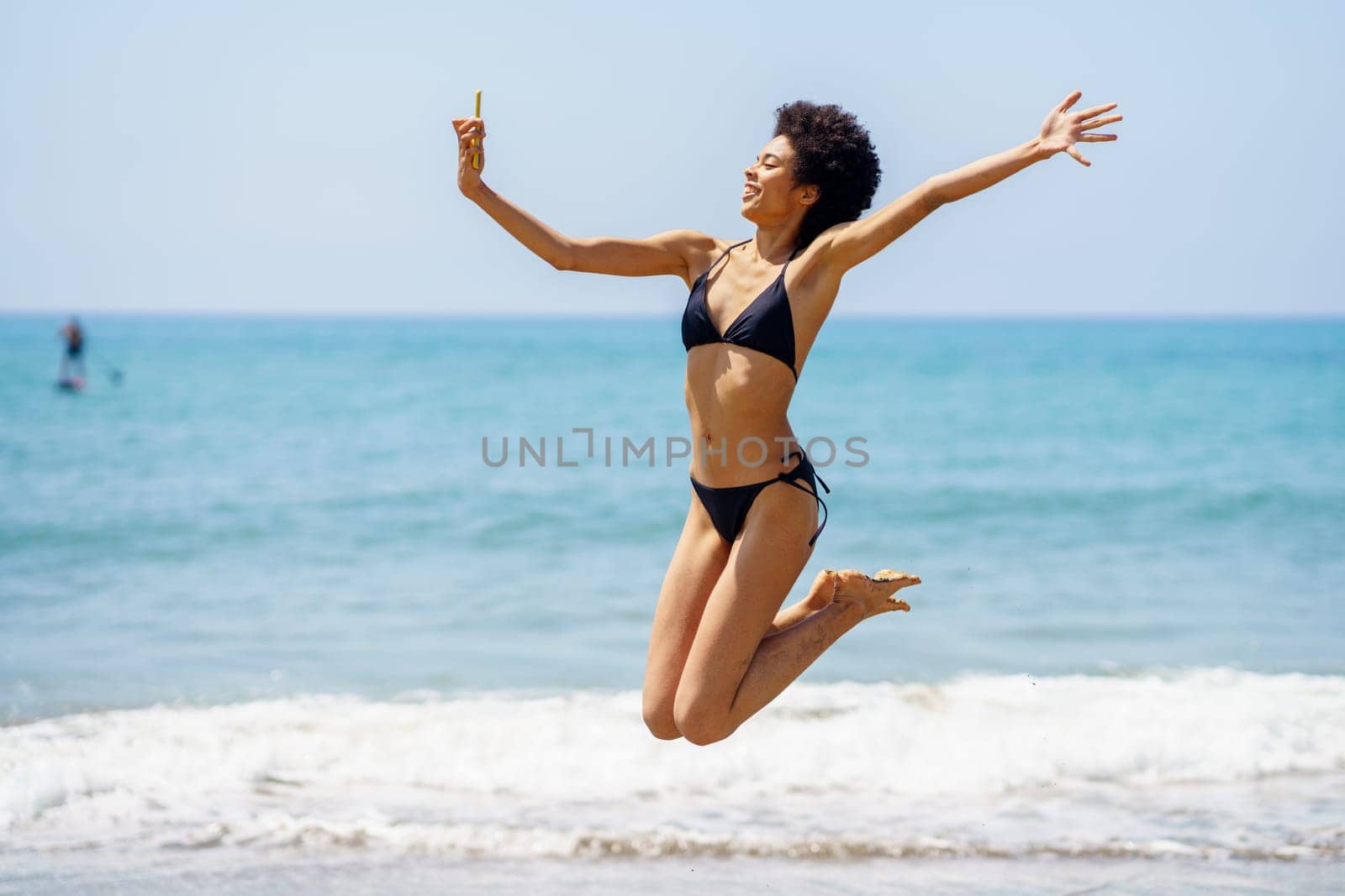 Full body side view of cheerful African American female traveler in bikini taking self portrait on smartphone and jumping on coast near waving sea