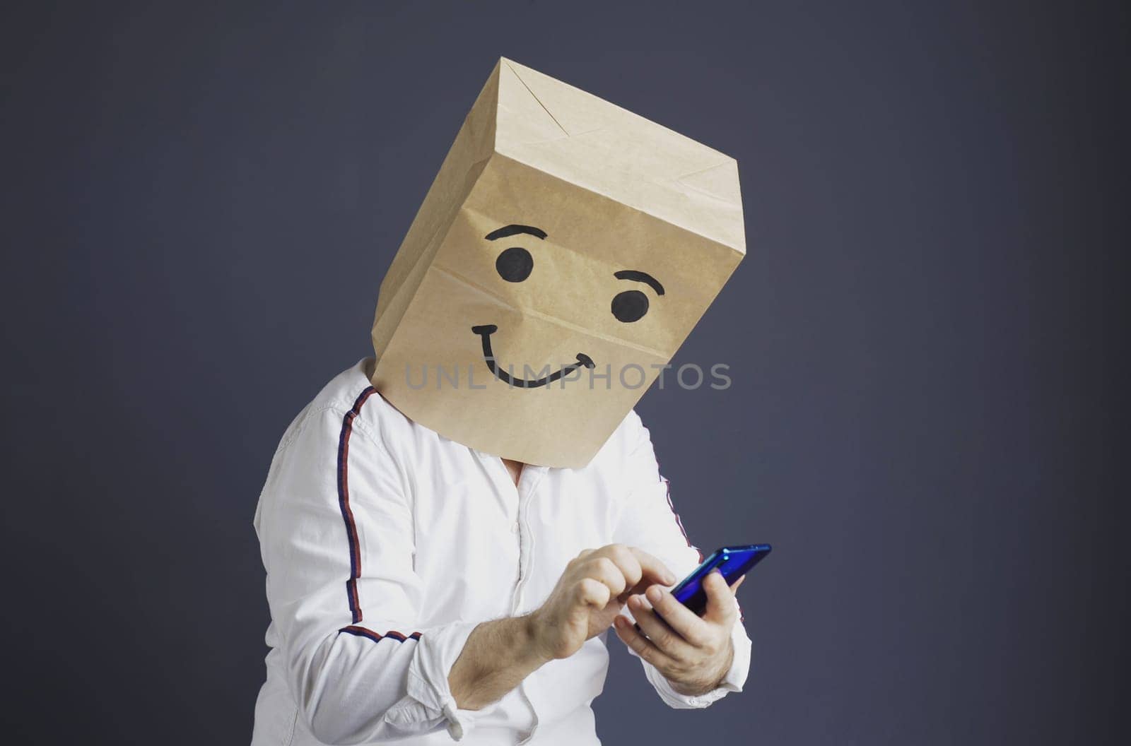 A man in a white shirt with a paper bag on his head, with a smiley face drawn, writes a message and takes a selfie on a smartphone.