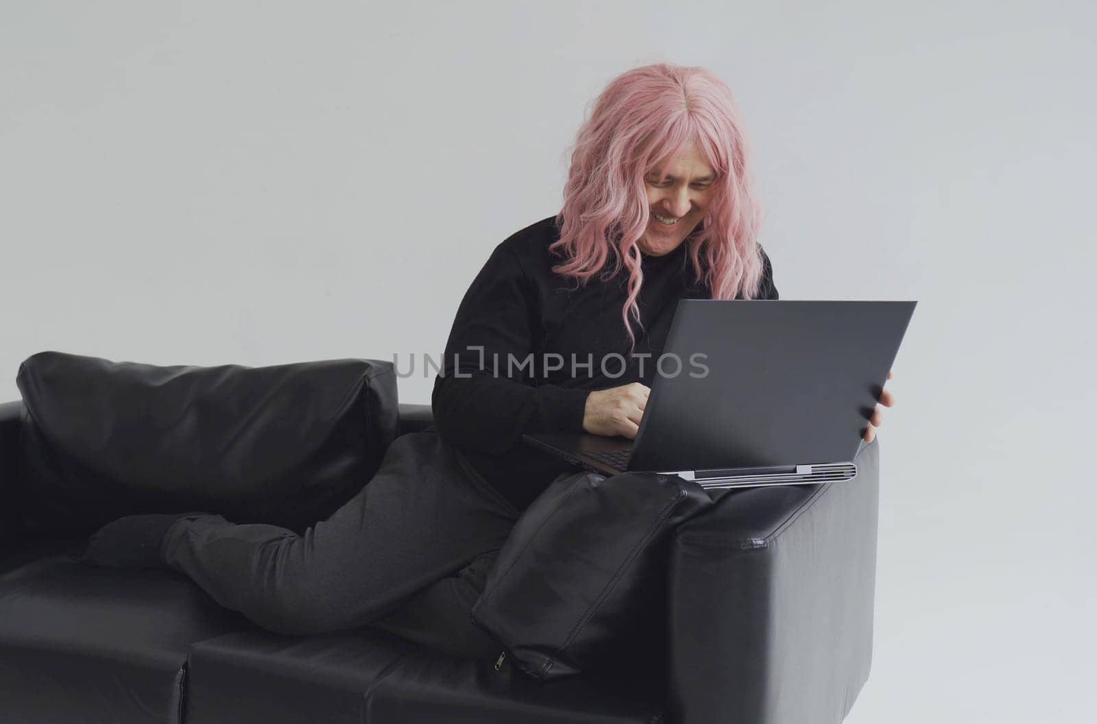 Portrait of a man in a pink wig, sitting on a sofa, typing on a laptop. White background.