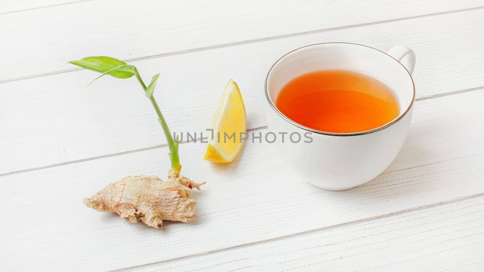 White cup with hot amber tea, lemon and dry ginger root with green spring next to it, on white boards. by Ivanko
