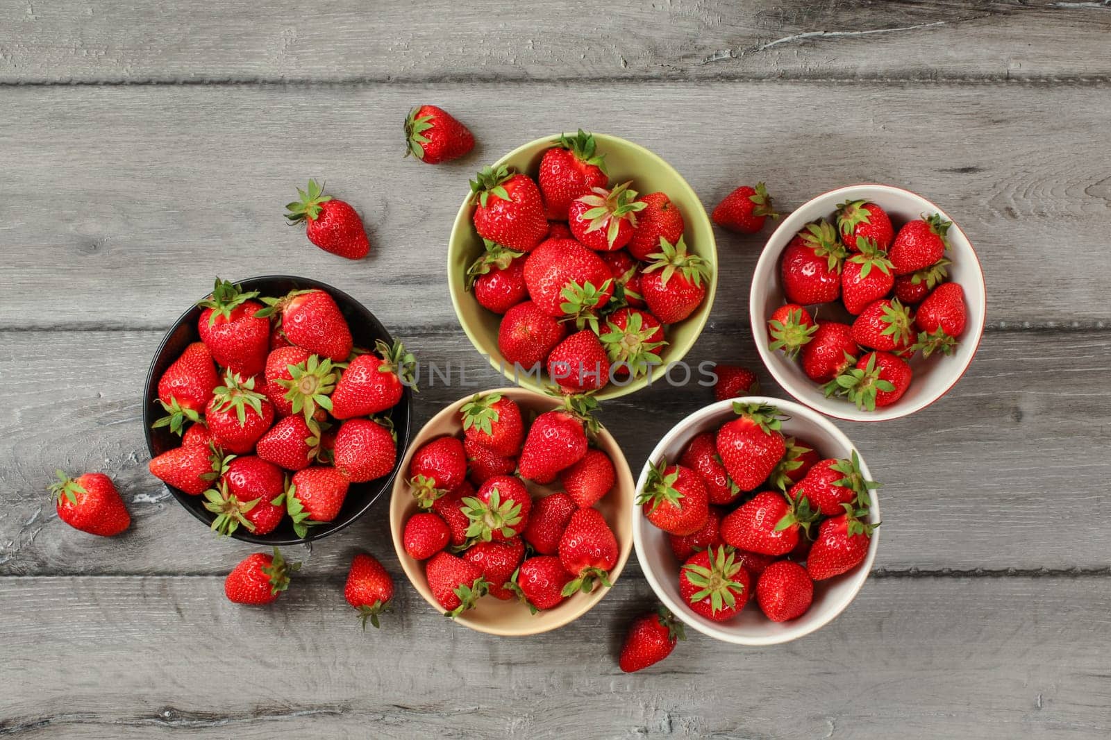 Tabletop view - five small bowls with strawberries, some of them spilled on gray wood desk.