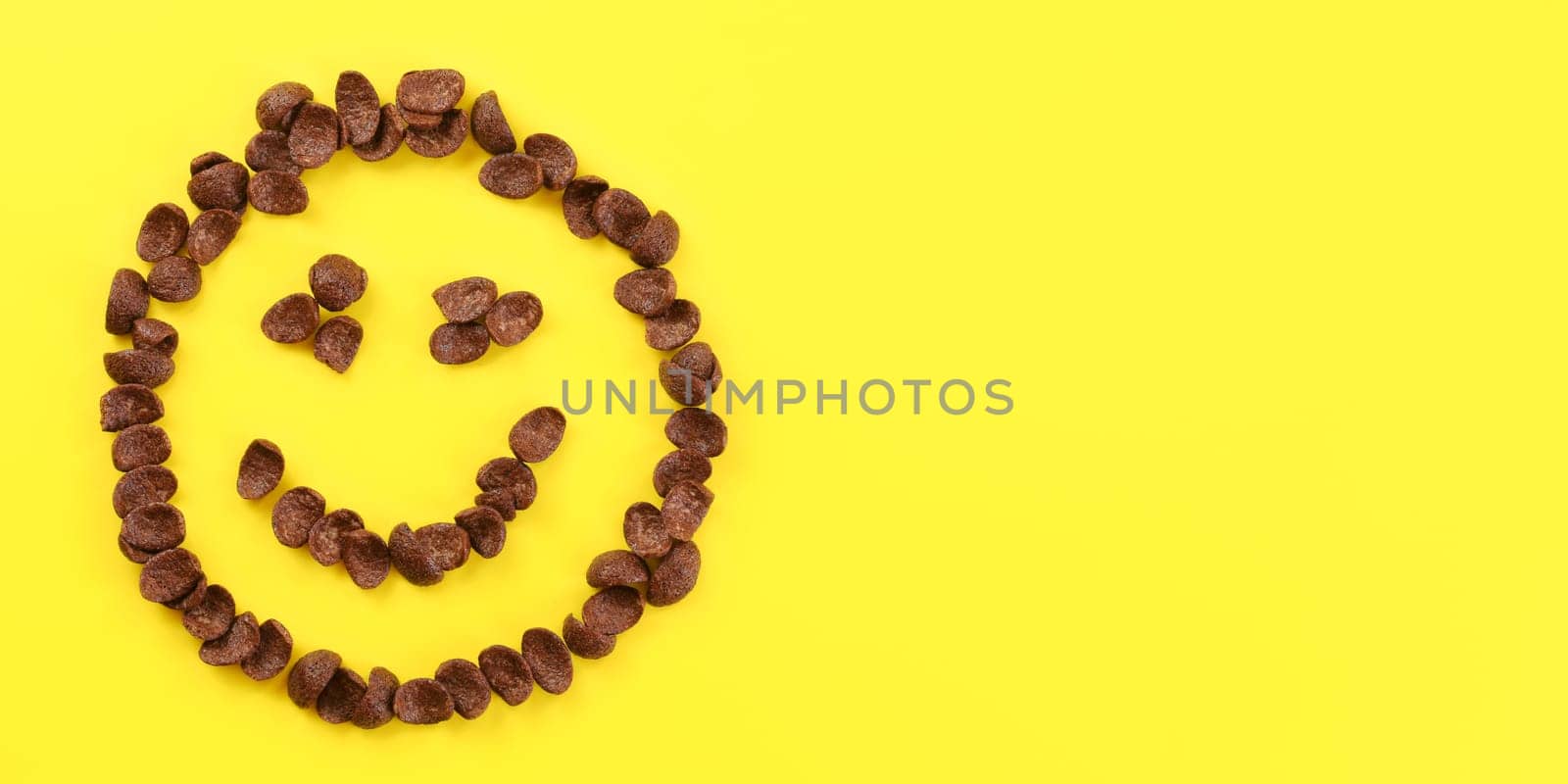 Tabletop view, smiley face made of brow chocolate corn flakes on yellow board. Space for text on right side.