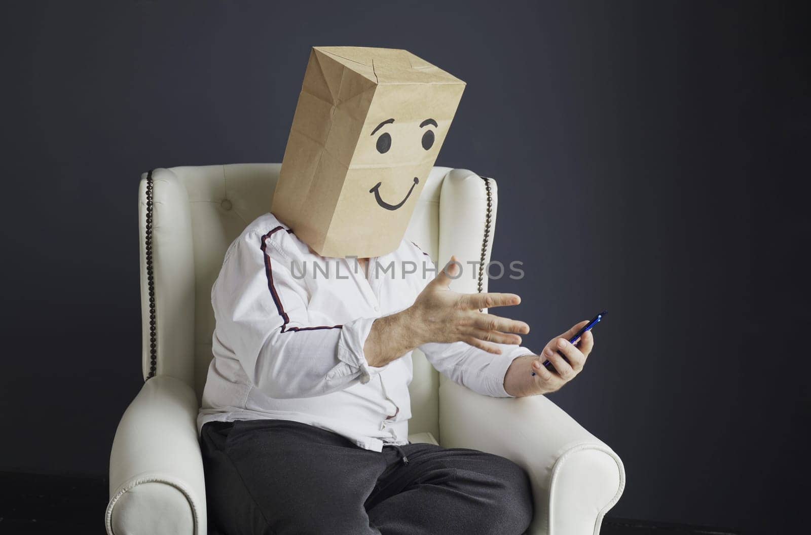 A man in a white shirt with a paper bag on his head, with a drawn smiling emoticon, is talking on a video call on a smartphone, gesturing with his hand.