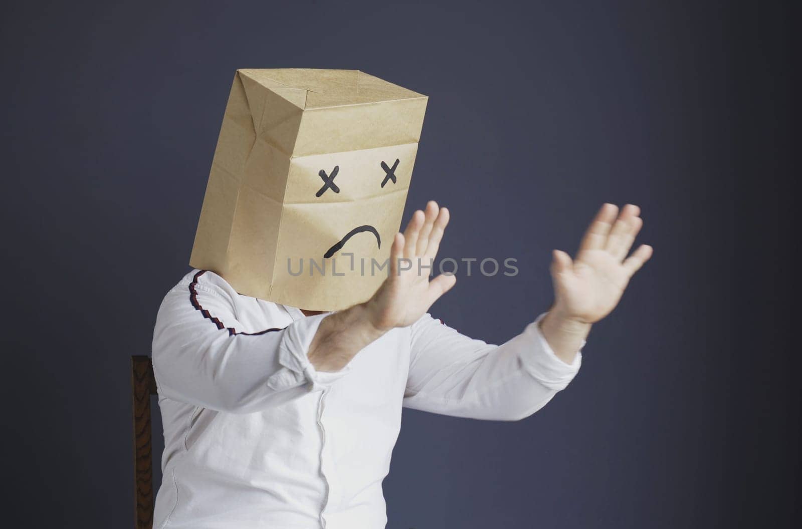 A sad man in a white shirt with a bag on his head, with a sad emoticon drawn, is afraid, defends himself by putting his hands forward. Emotions and gestures.