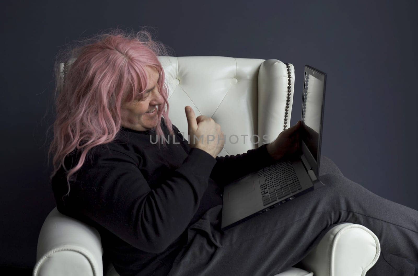 A man in a pink wig sits in a white chair and communicates via video link on a laptop. Side view