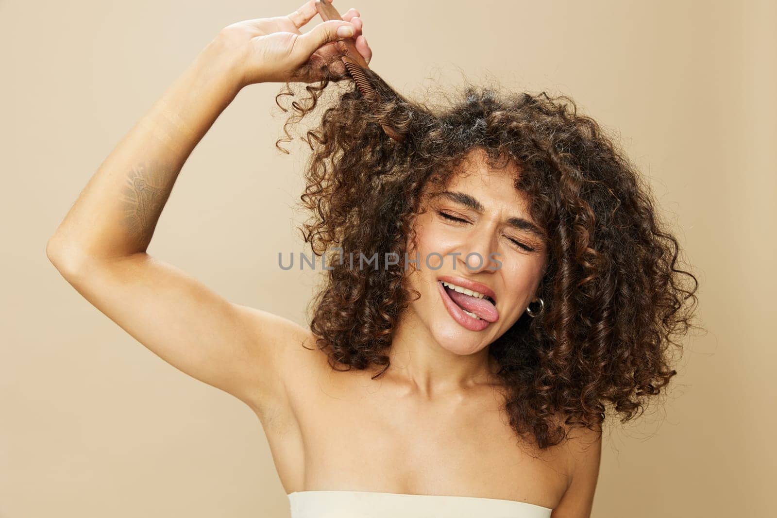 Woman combs curly afro hair with wooden comb, home beauty care styling products, hair loss after covid-19, smile on beige background. High quality photo