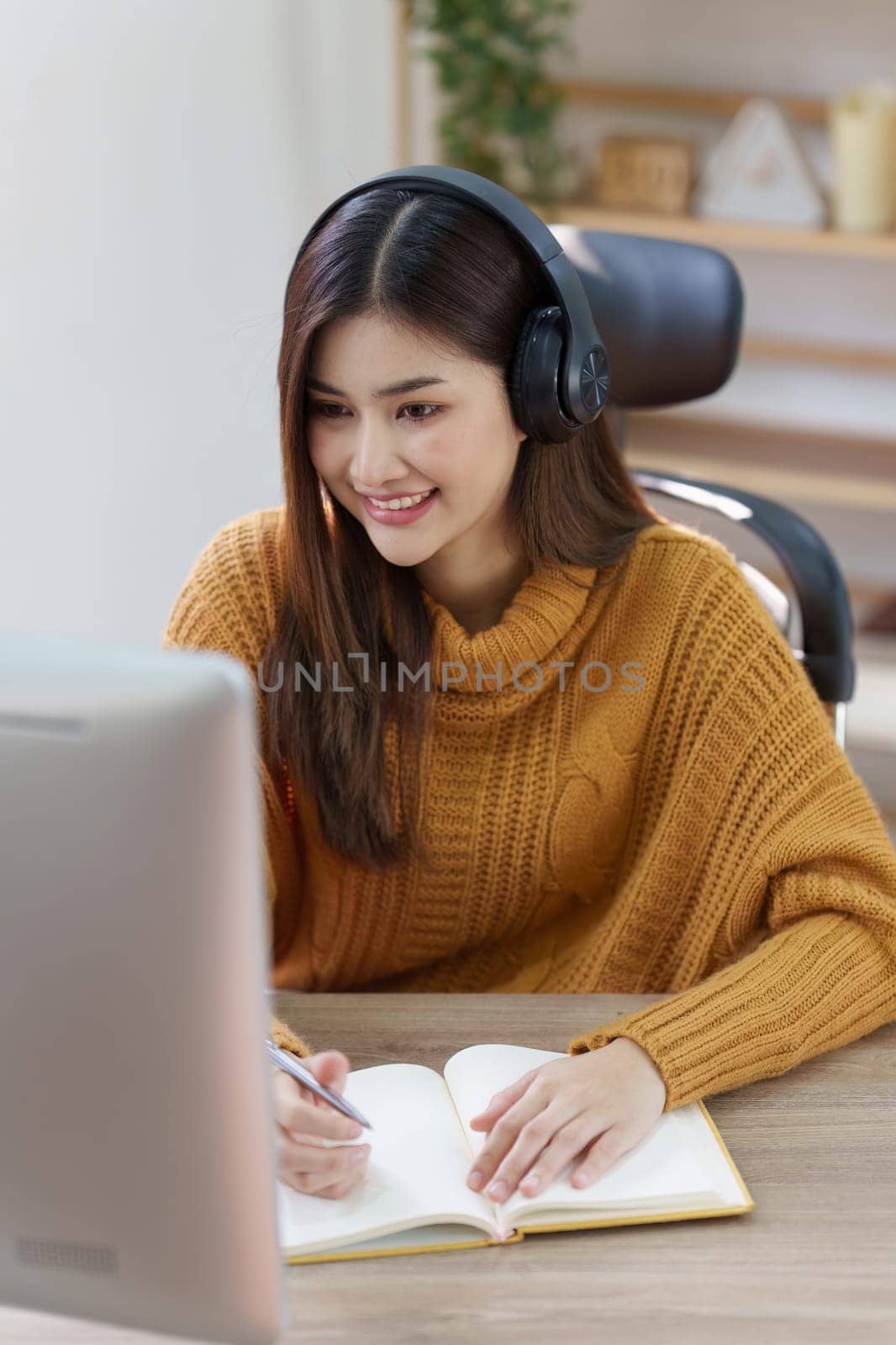 Online education, e-learning. Asian woman in stylish casual clothes, studying using a computer, listening to online lecture, taking notes, online study at home.